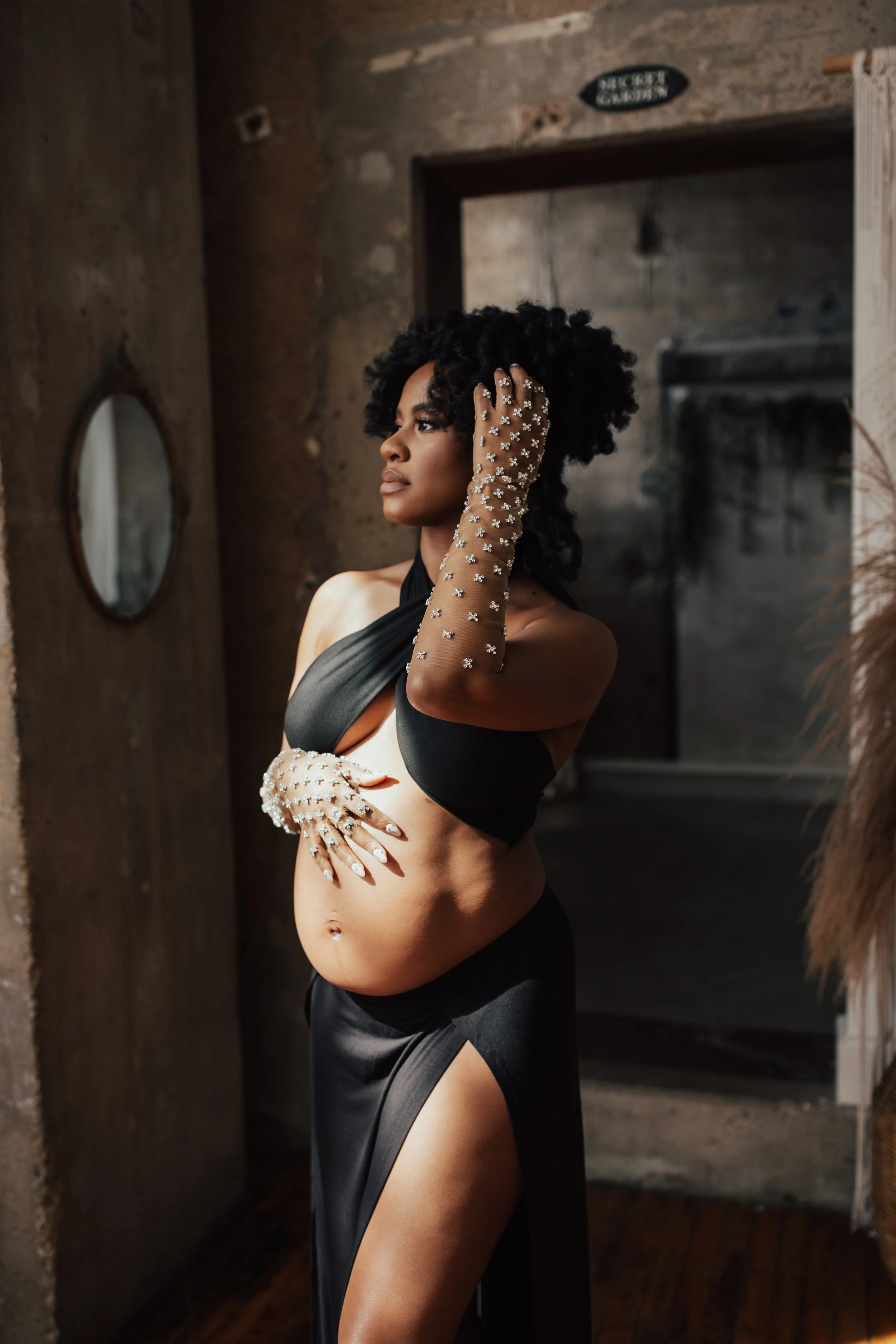 A woman with curly black hair stands wearing a black halter top and a black skirt with a high slit. She is adorned with sheer gloves and matching nail jewelry covered in pearls and rhinestones. Dallas Fort Worth DFW Texas maternity photographer