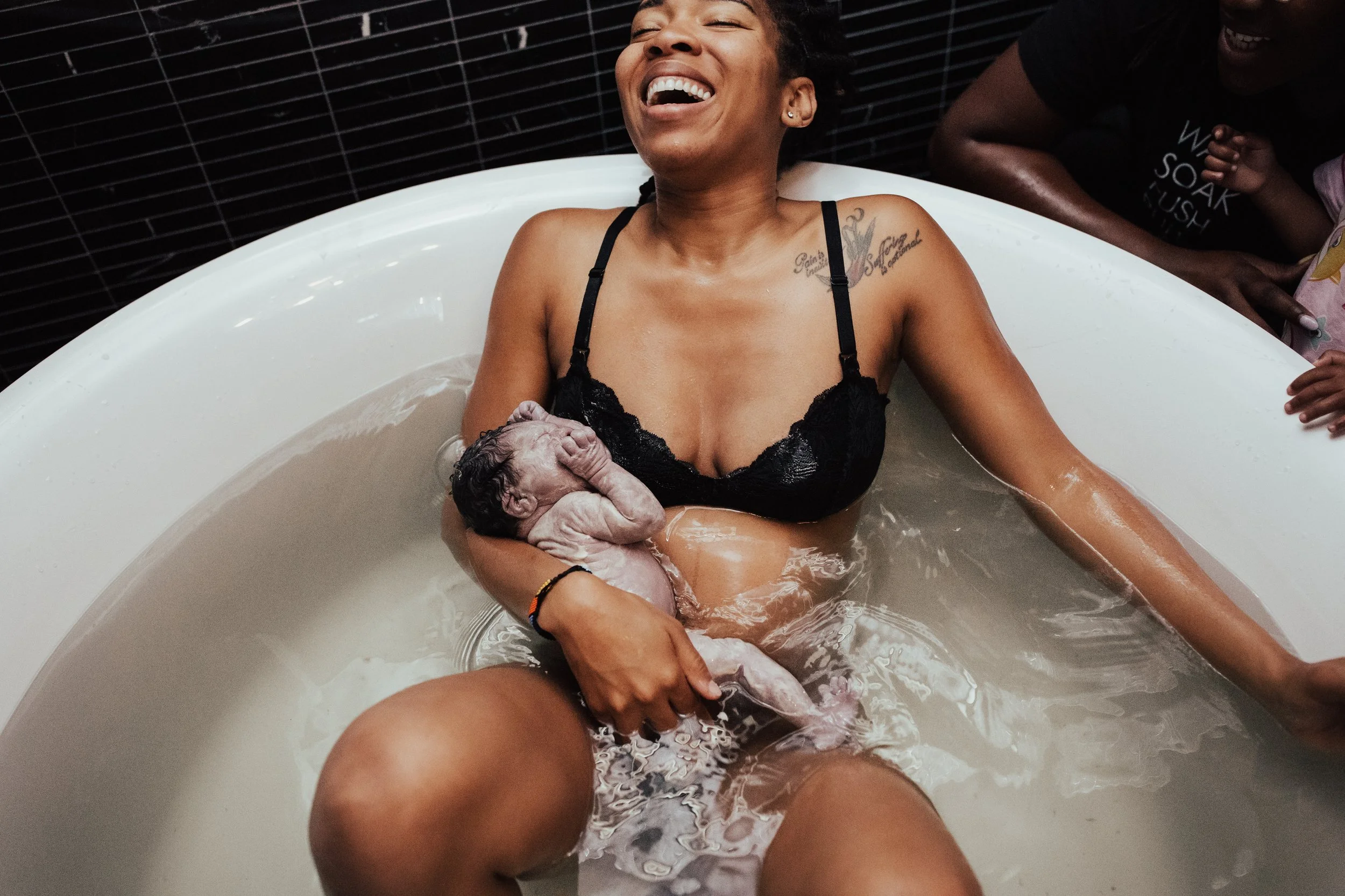 A woman in a bathtub holding a newborn baby, smiling and laughing, with two children watching nearby.
