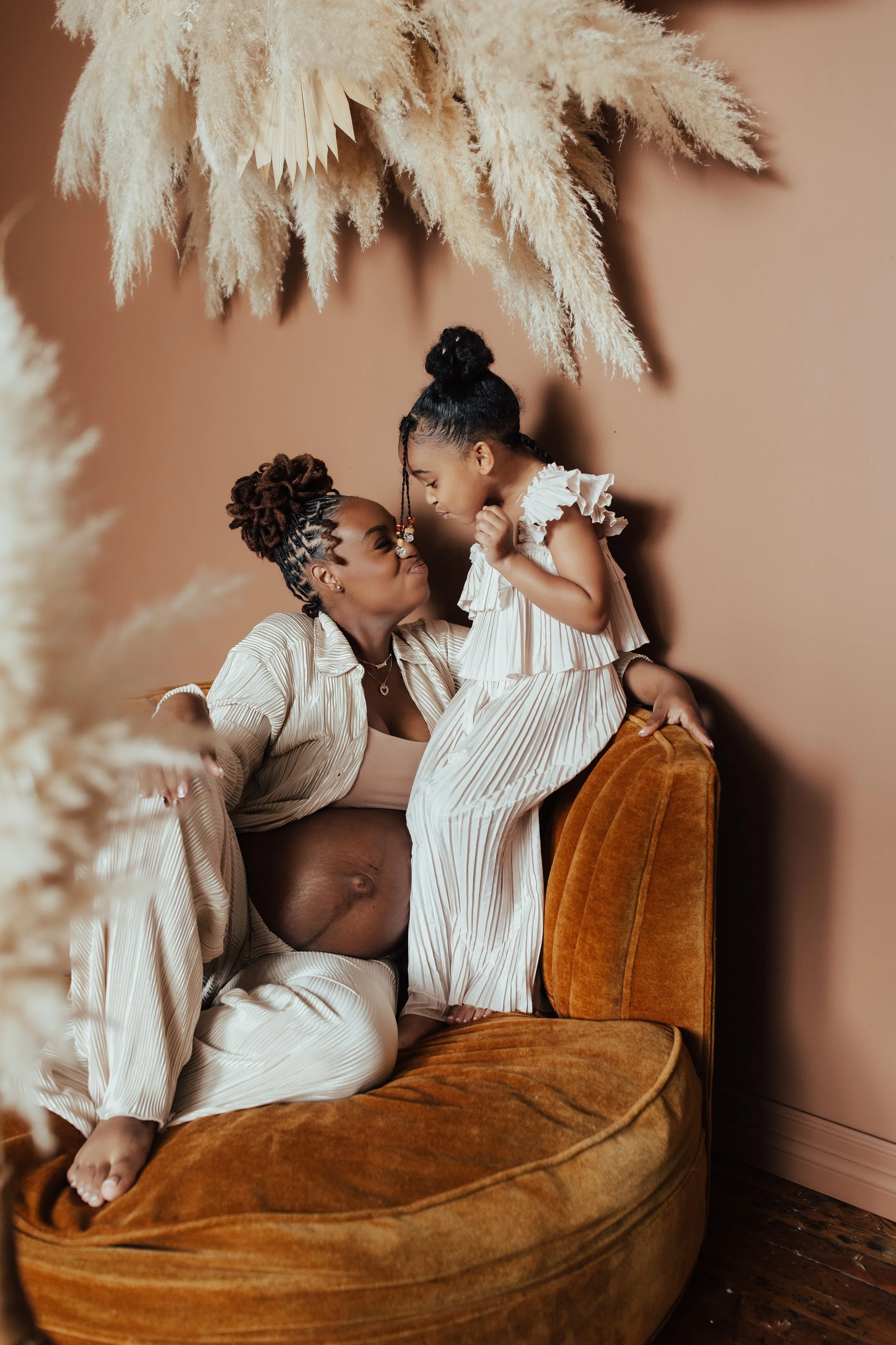 A woman with a pregnant belly and wearing pajamas is sitting on a vintage orange sofa, interacting affectionately with a young girl who is standing on the sofa. Dallas Fort Worth DFW Texas Maternity Photographer