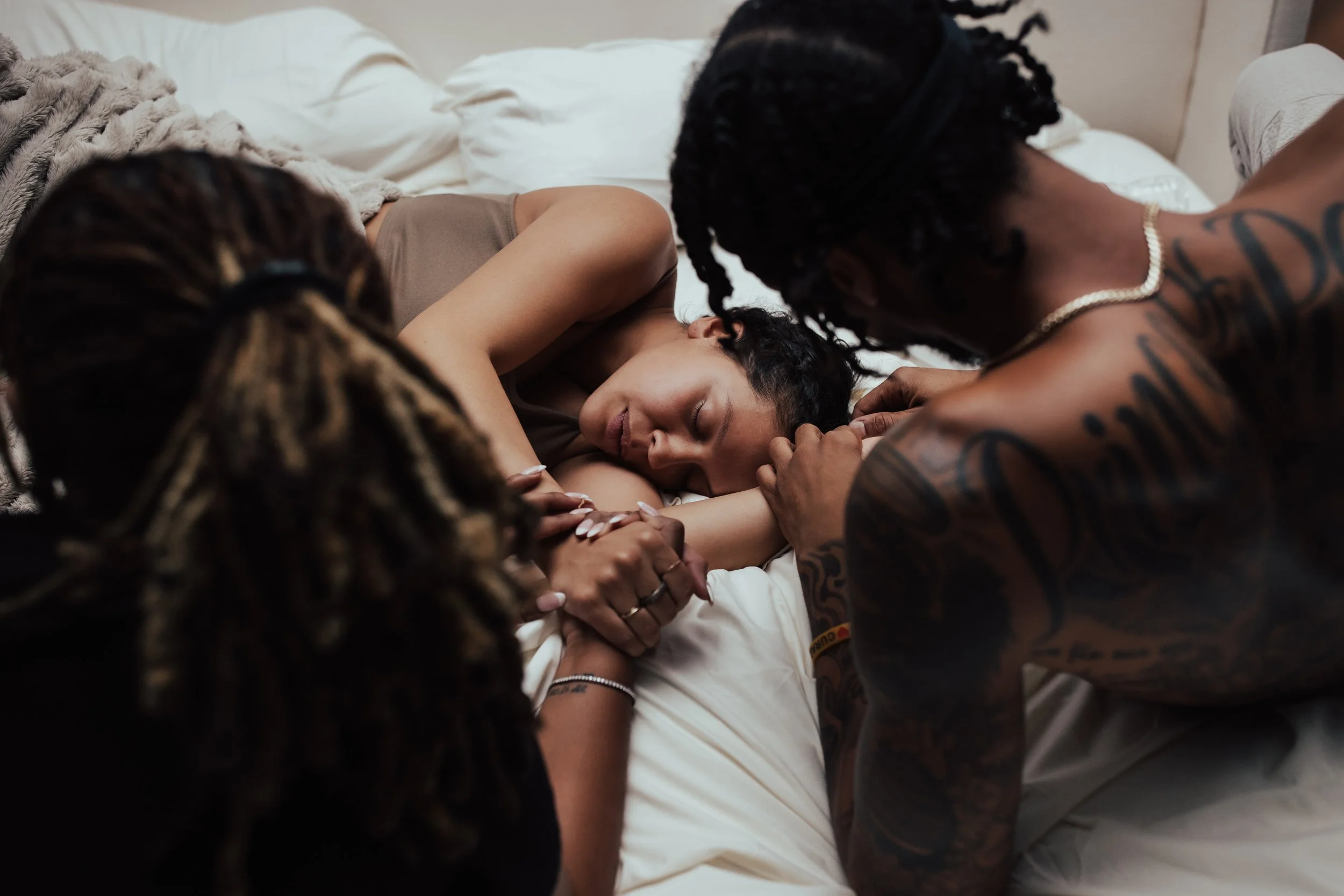 Three women with dreadlocks and tattoos lying on a bed, cuddling and holding hands. One woman is resting with her eyes closed, seemingly relaxed or sleeping.