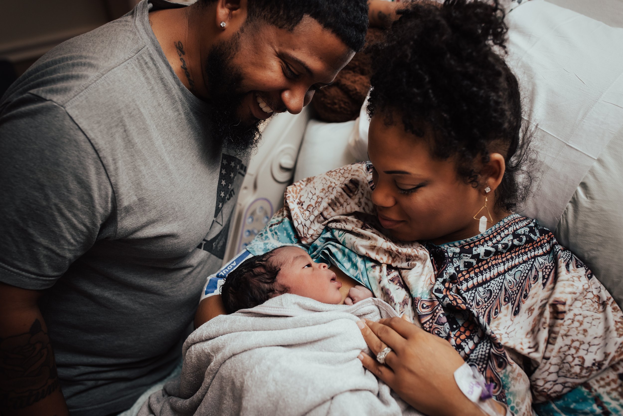 New mother lying in a hospital bed, holding her newborn baby wrapped in a blanket, while her partner leans over smiling at them in a hospital room.