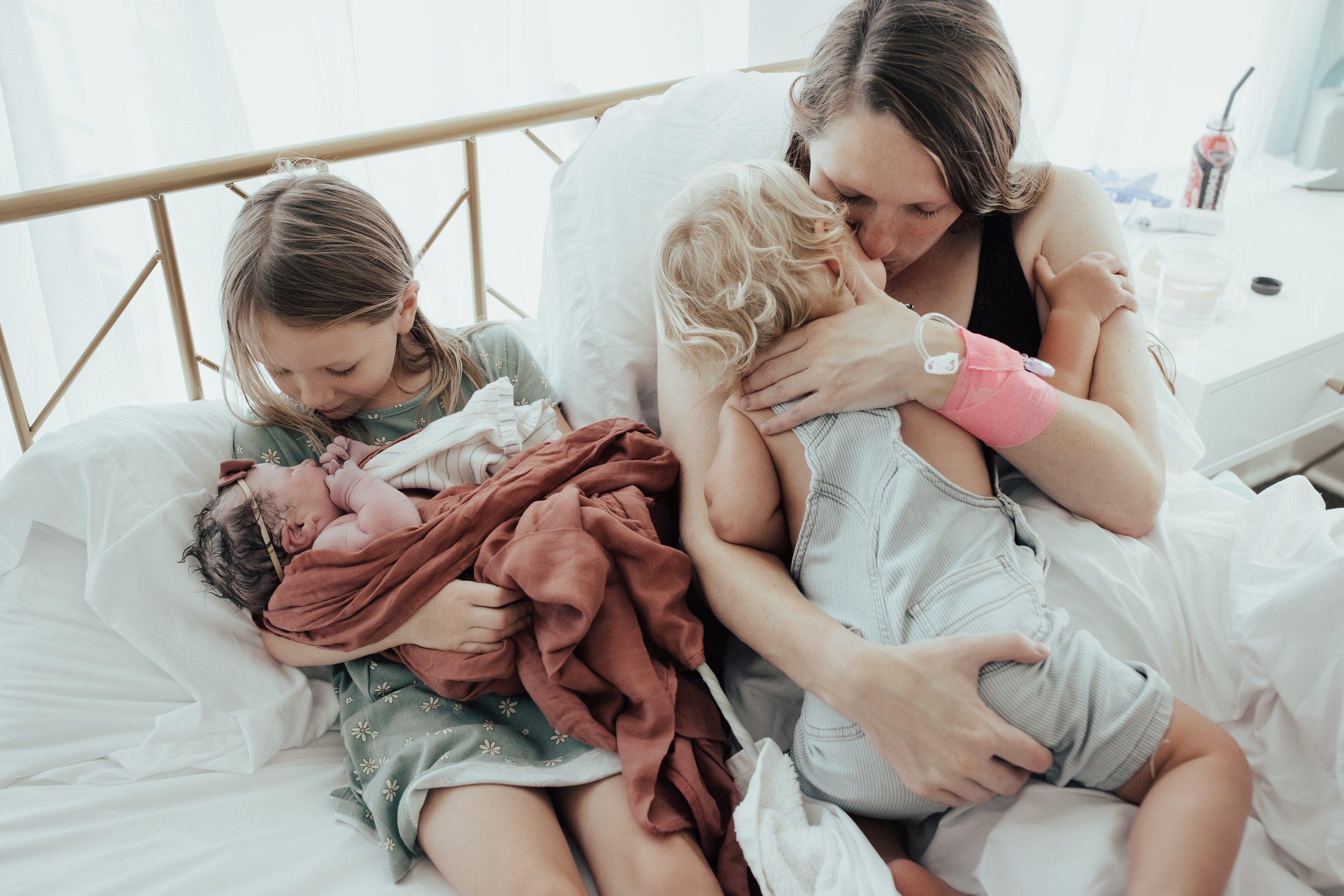 A woman with two children in a hospital room post childbirth, holding a newborn wrapped in a blanket, and interacting with a young child who is also holding a newborn.