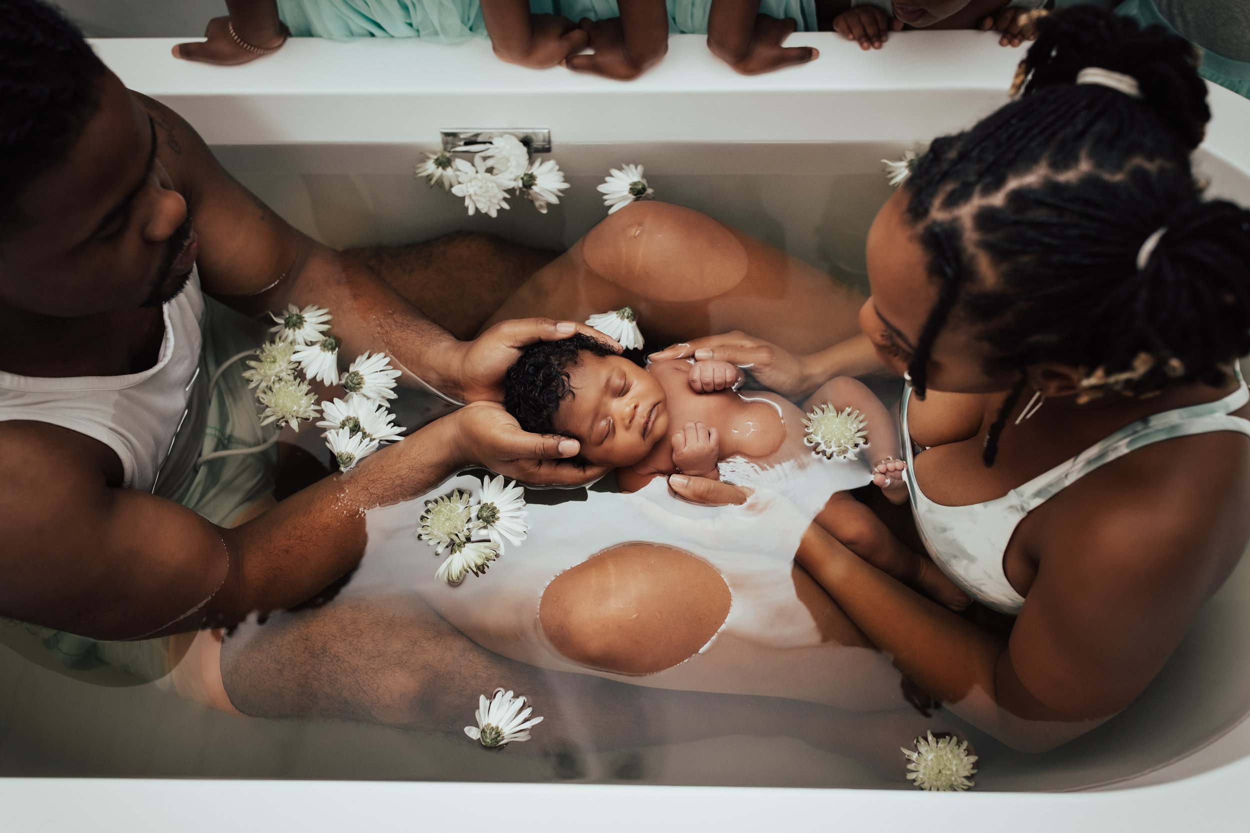 A newborn baby is taking a bath in a bathtub surrounded by two adults with flowers floating in the water.