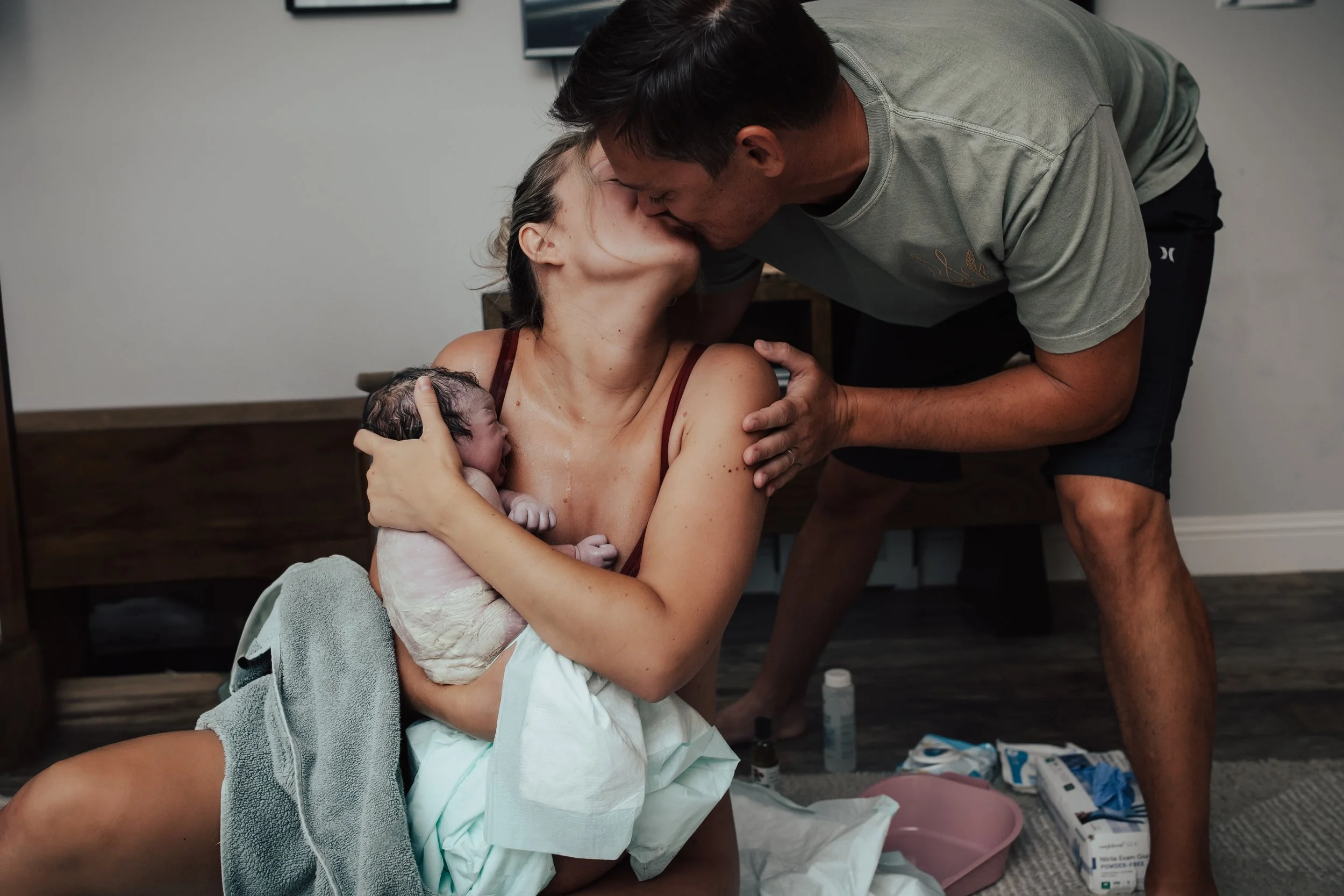 Woman holding a newborn baby after birth, supported by a towel, as a man leans over to kiss her in a hospital room. Dallas Fort Worth Maternity photoshoot Carmen Bridgewater photography