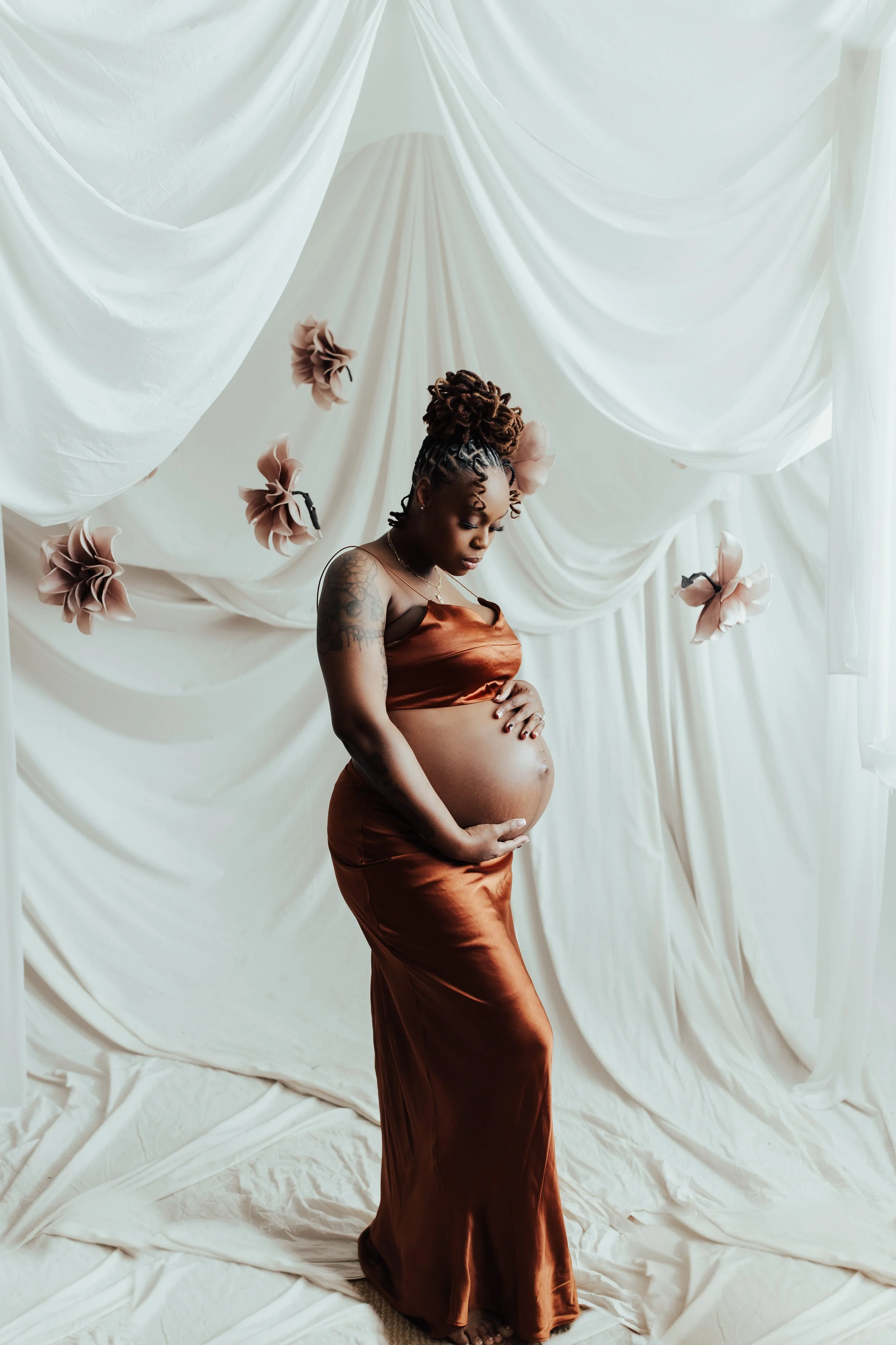 A pregnant woman with dark skin and styled hair in a high bun, wearing a satin copper-colored dress, standing in front of a cream-colored draped fabric backdrop with floating soft pink flowers. Dallas Fort Worth DFW Texas Maternity Photographer
