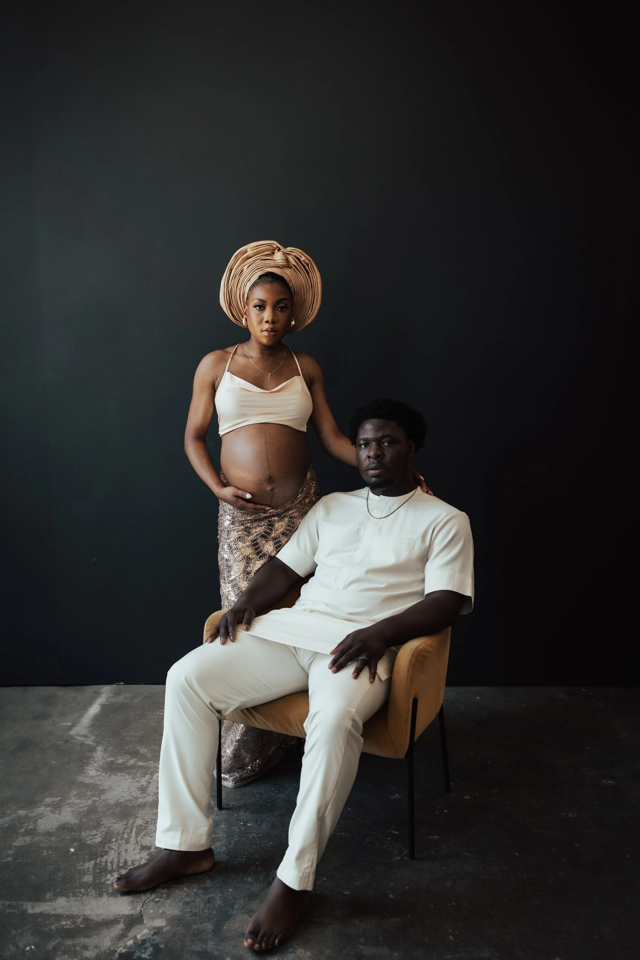 A pregnant woman standing next to a seated man, both dressed in elegant white and beige attire, posing against a dark studio background. Dallas Fort Worth DFW Texas Maternity Photographer