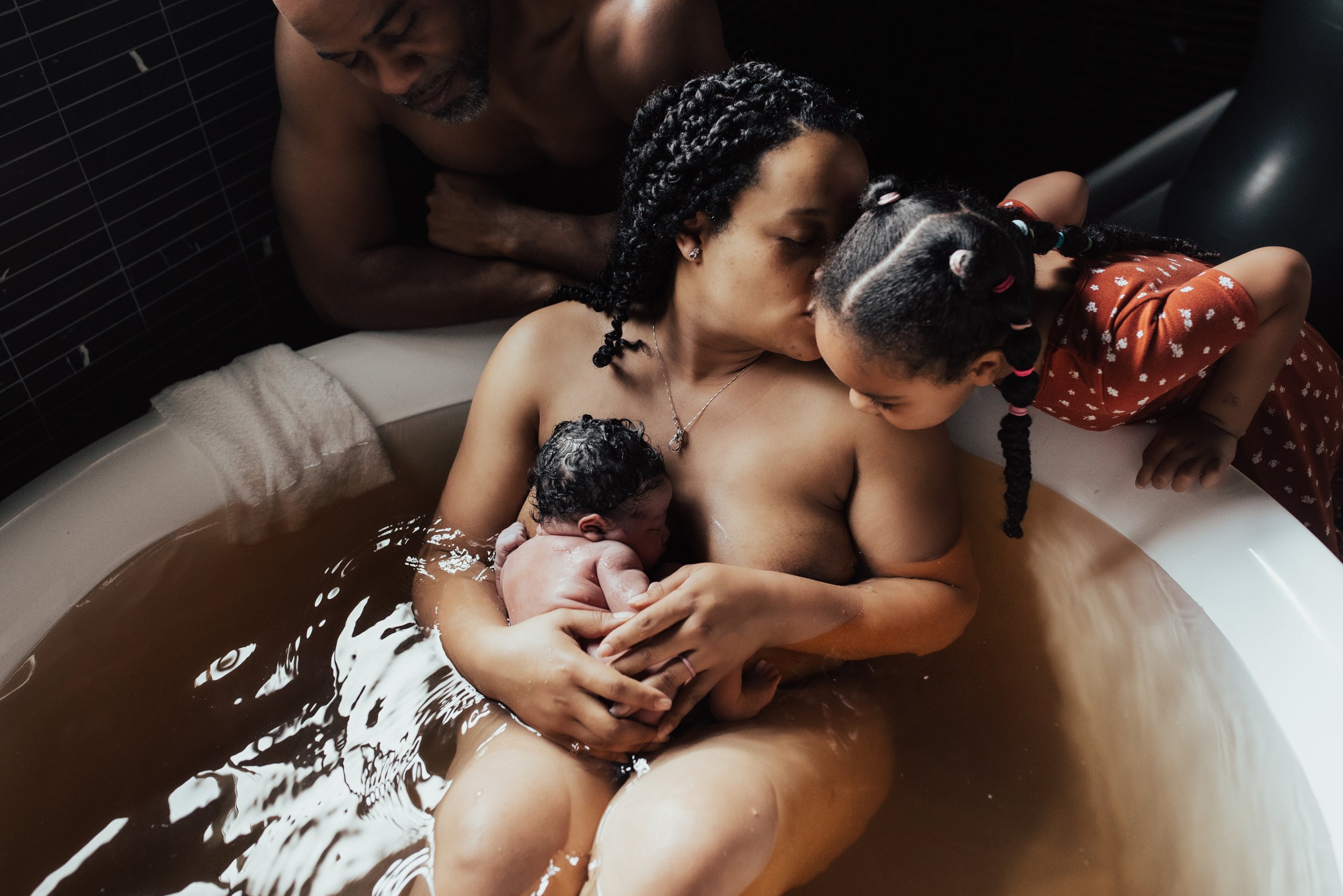 Family in a bath, a woman with a newborn, a young girl leaning over kissing the woman, a man watching in the background. Dallas Fort Worth Maternity photoshoot Carmen Bridgewater photography