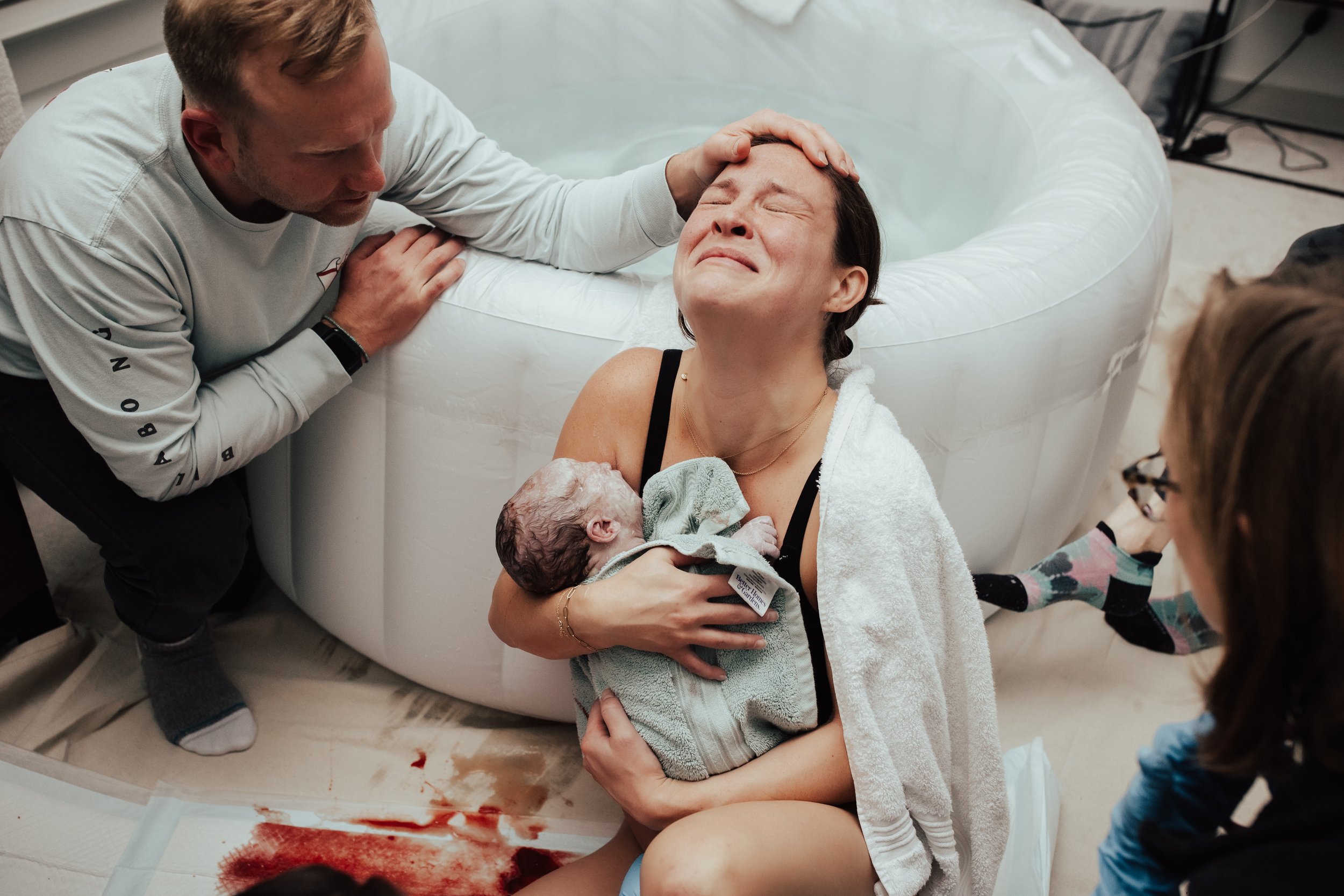New mother holding her newborn baby, surrounded by supportive family members at a water birth in a birthing pool, with a towel draped over her shoulder and expressive emotion on her face.