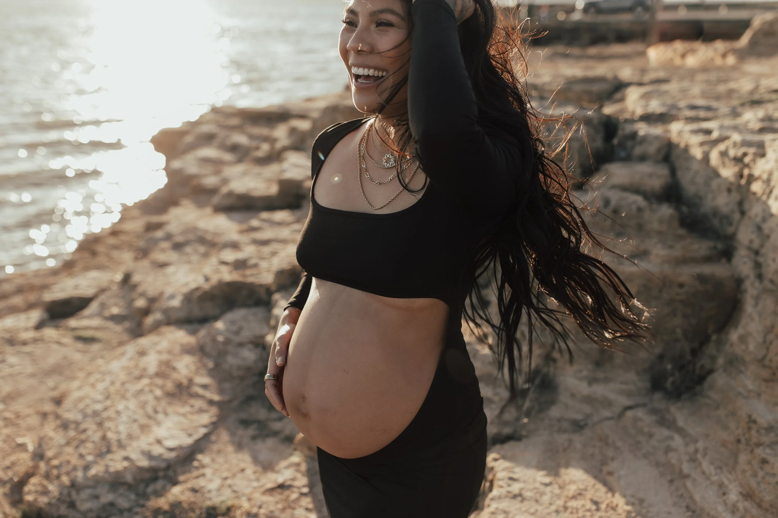 A pregnant woman with long dark hair, wearing a black crop top and jewelry, smiling and standing on a rocky shoreline near water during sunset.