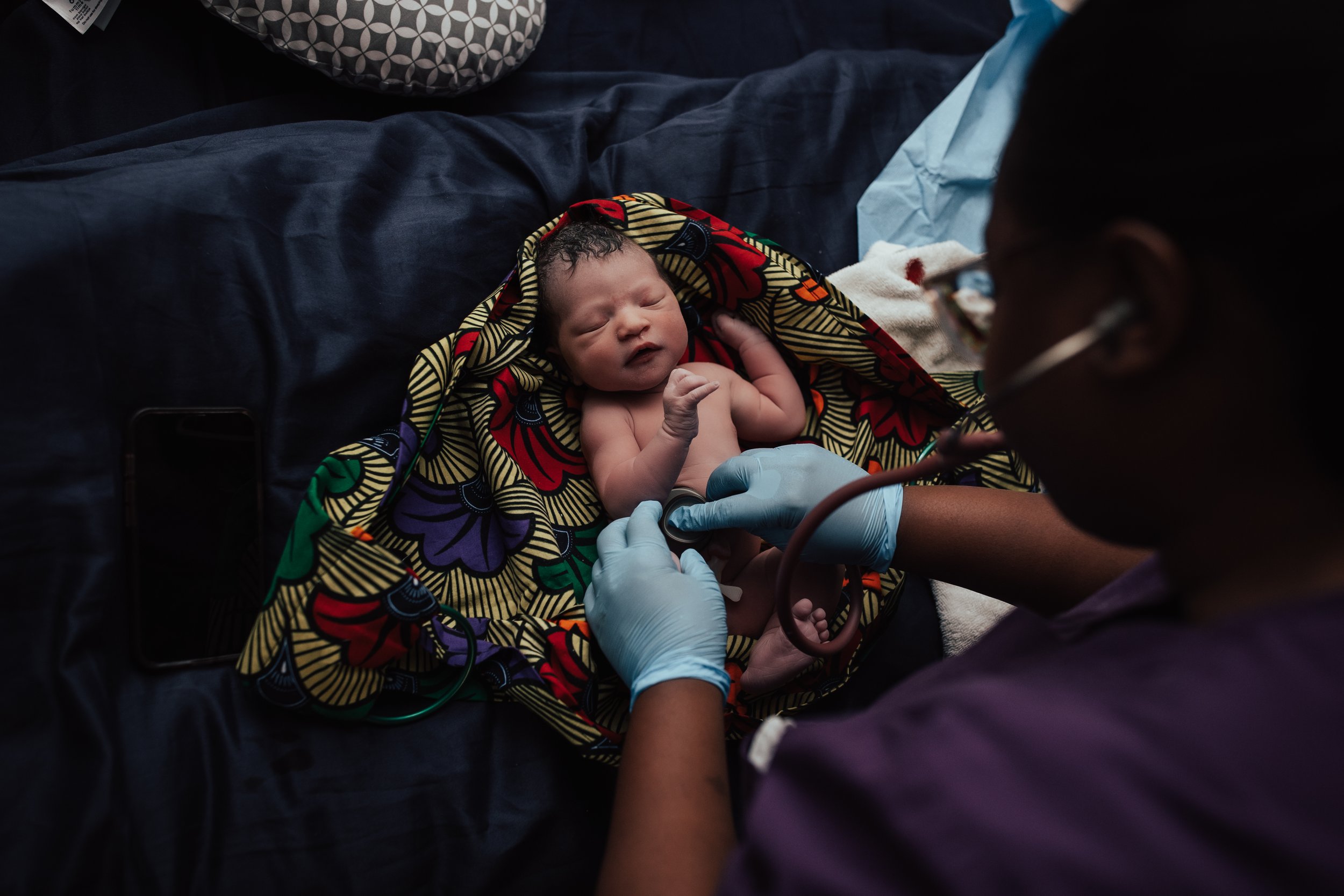 A newborn baby lying on a colorful blanket, with a Dallas midwife worker in scrubs and gloves examining the baby using a medical stethoscope. Dallas Fort Worth Maternity photoshoot Carmen Bridgewater photography