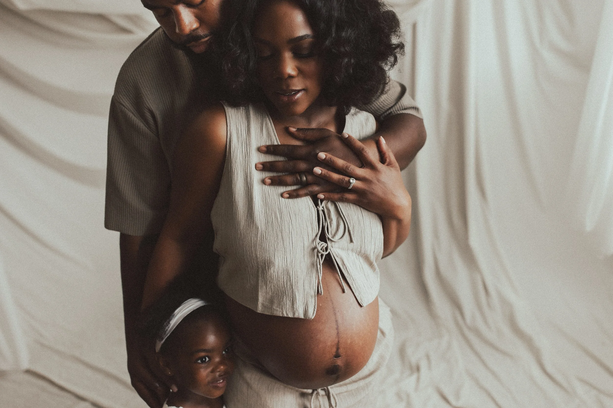 A pregnant woman with dark skin and curly hair, wearing a light-colored top, stands in front of a backdrop with beige drapes. Dallas Fort Worth DFW Texas Maternity Photographer