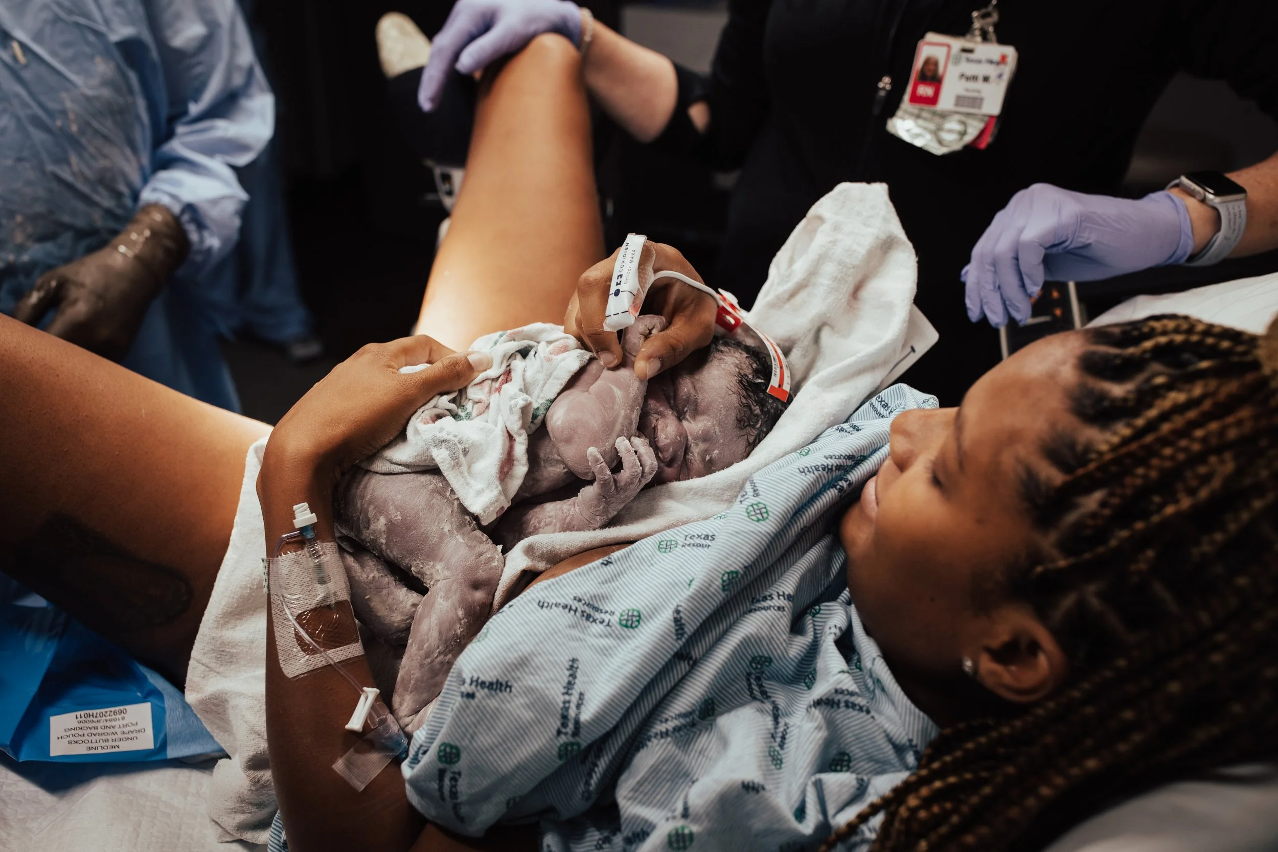 A woman in a hospital bed holds her newborn baby, who is covered in vernix and still in the amniotic sac. Medical staff surround the woman, assisting with the delivery.