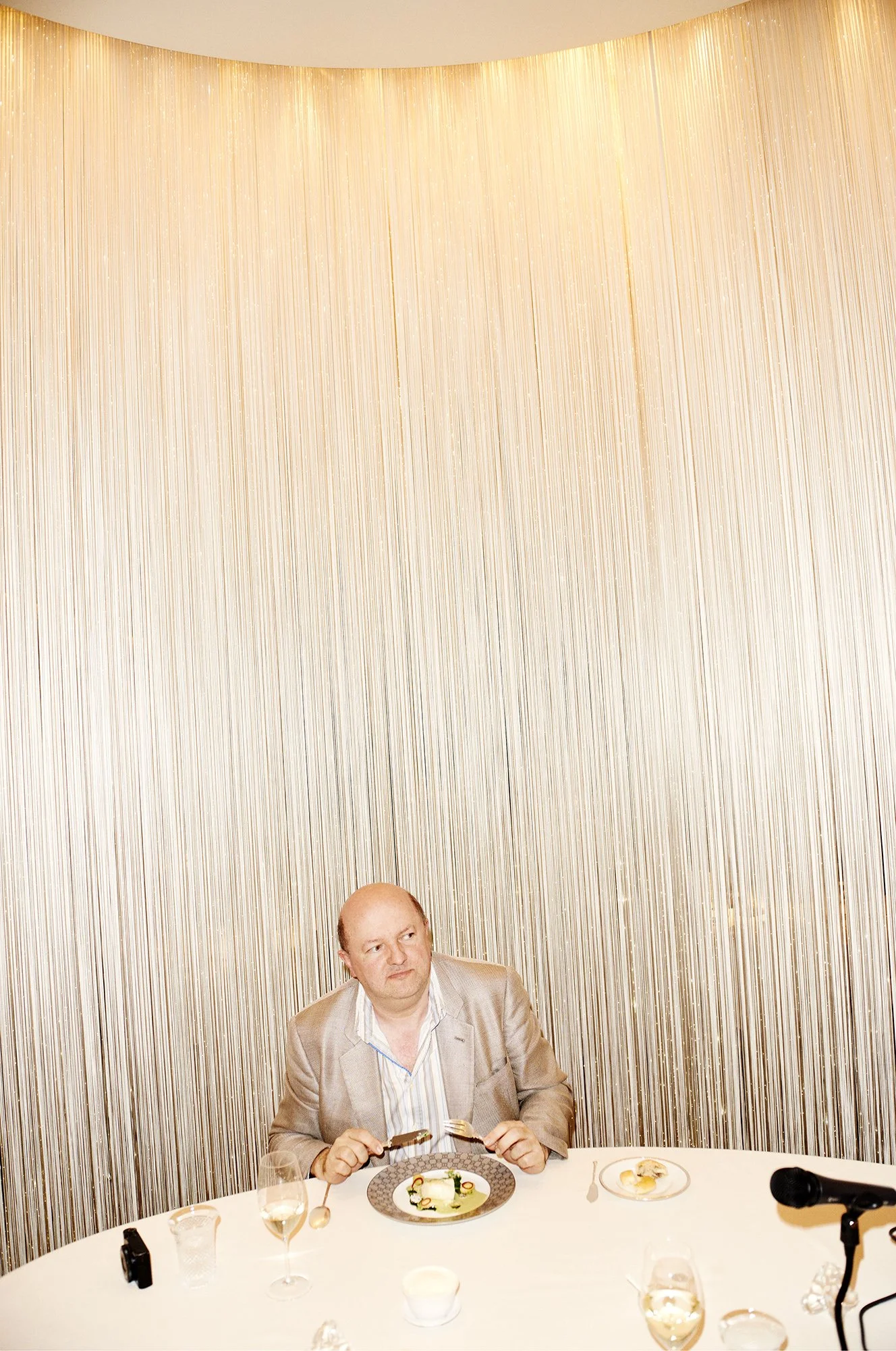A man in a beige suit sitting at a round table with a bottle of water, glasses, and a plate with food, holding utensils, in a room with a large, patterned wallpapered wall behind him.