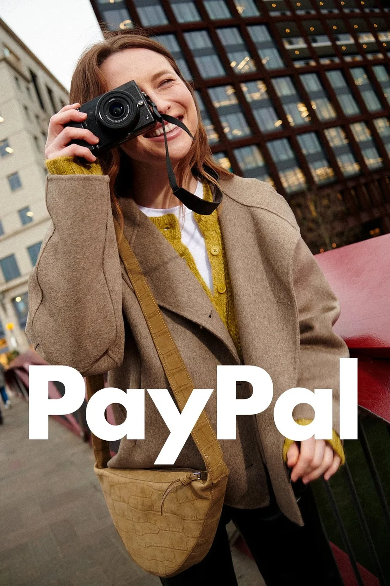 Woman smiling and holding a camera outdoors in an urban setting with a downtown building and street in the background, with the PayPal logo overlayed.