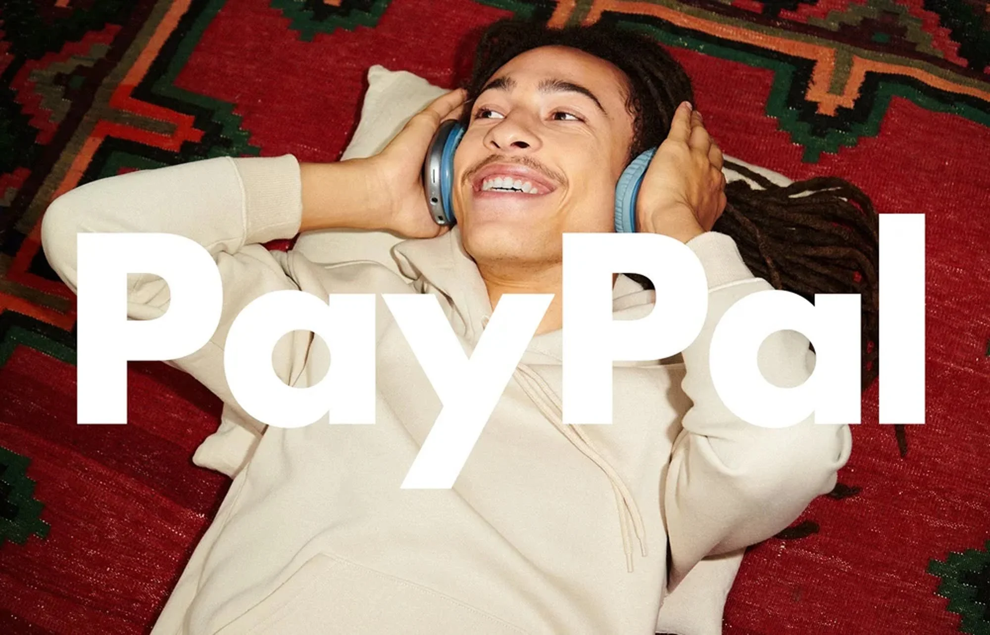 Person lying on a colorful red and green patterned rug, smiling, with eyes closed, wearing a beige hoodie and listening to music with blue headphones.