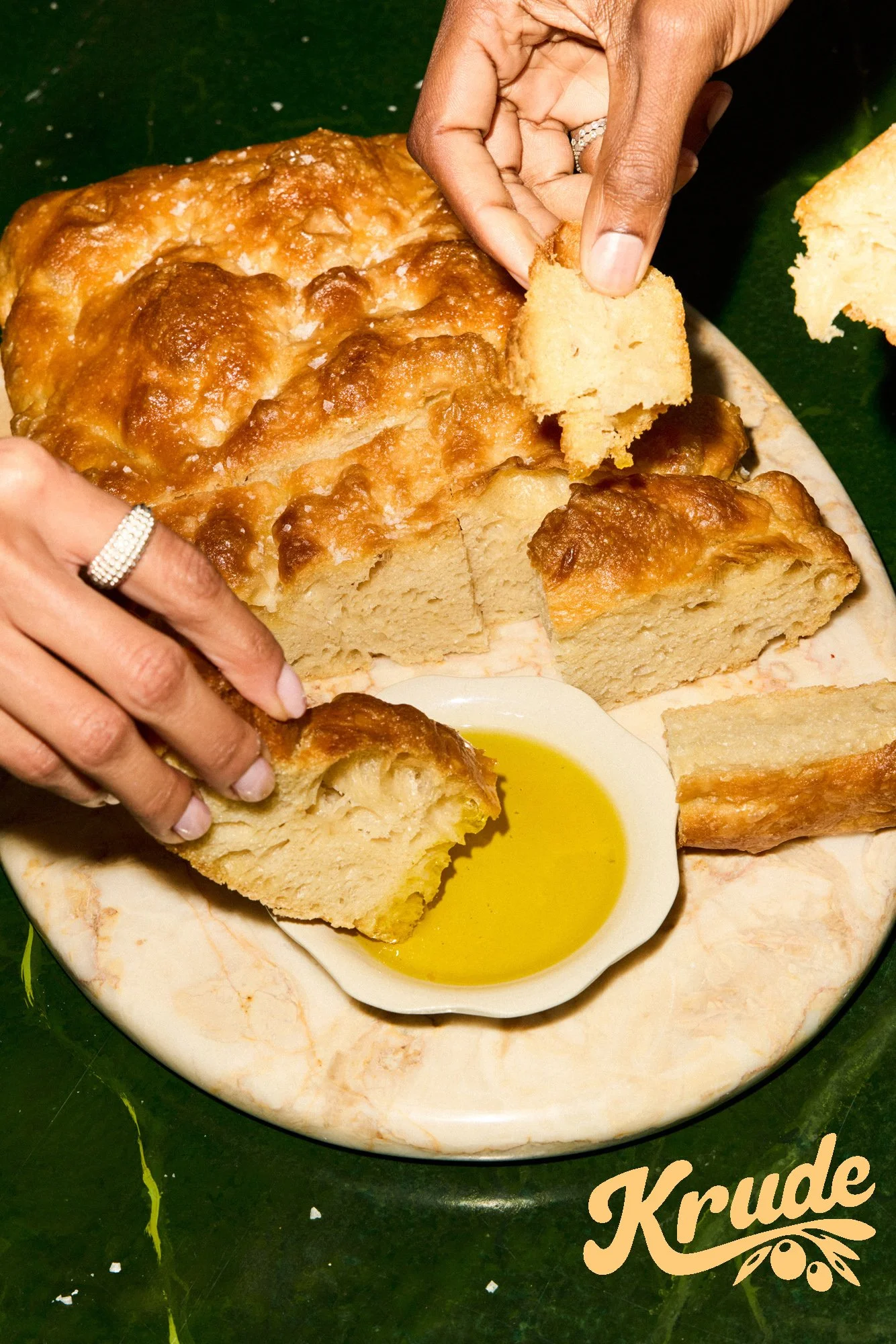 Close-up of a person's hands breaking apart a large piece of freshly baked cornbread on a marble serving platter, with a small dish of melted butter or oil nearby. The platter is on a dark green surface with yellow decorative letters spelling 'Krude'