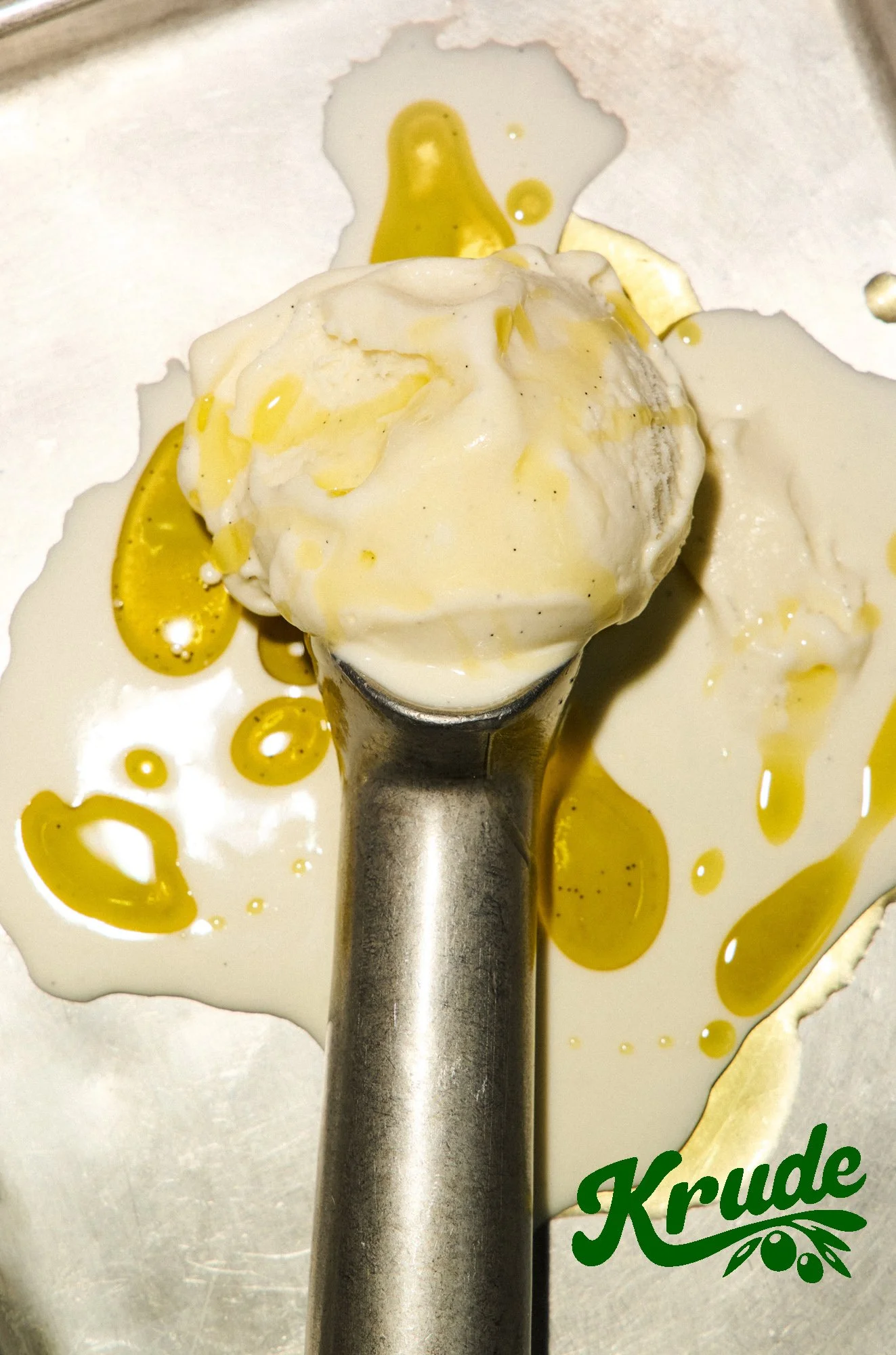 A scoop of vanilla ice cream with crumbled cookies, drizzled with yellow caramel sauce, on a metal ice cream scooper. The background shows a metallic surface and part of a logo reading "Krude" with olive branches.