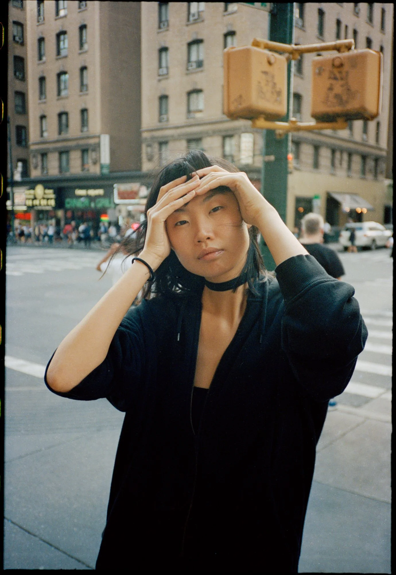 Young woman with black hair and choker necklace holding her head with both hands on a city street. Buildings, cars, and pedestrians in the background.