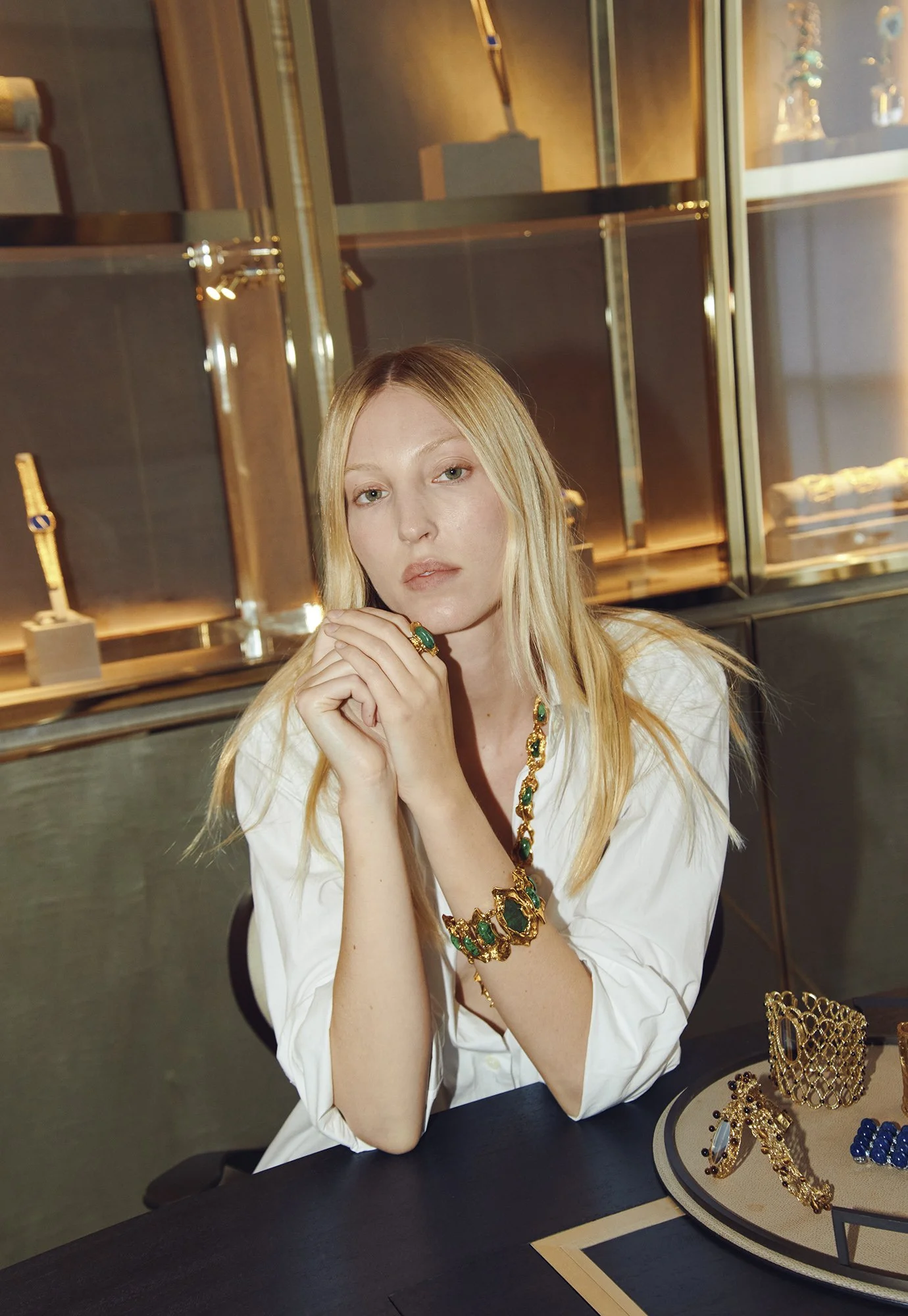 A woman with long blonde hair sitting at a table inside a jewelry store, wearing a white shirt and green and gold jewelry, including a ring, bracelet, necklace, and earrings. There are jewelry displays with gold and gemstone pieces on the table and i