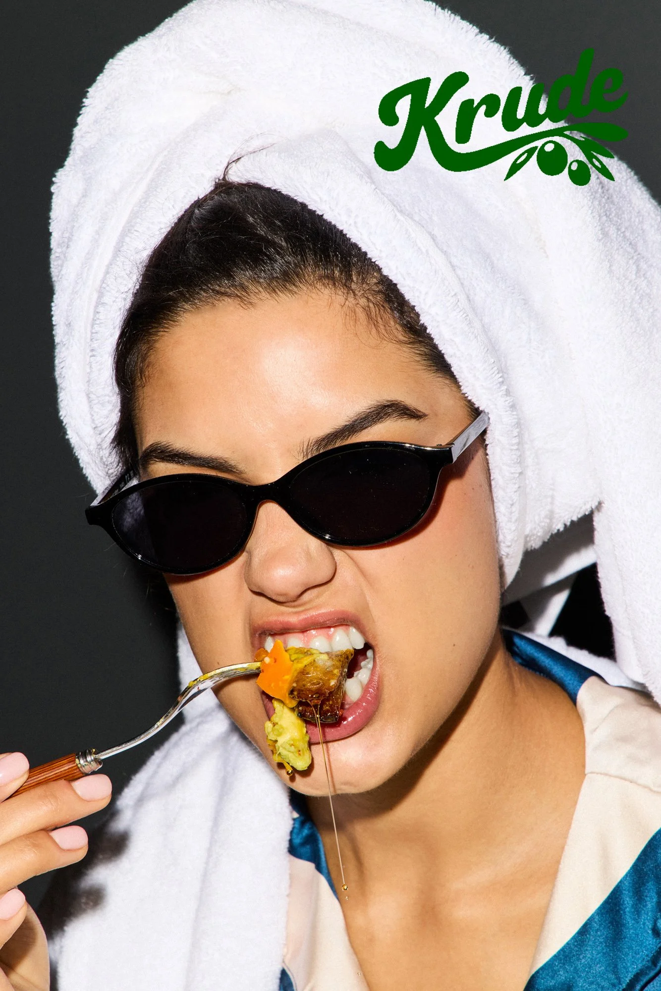 A woman with dark hair, wearing sunglasses and a white towel on her head, is eating food from a fork.