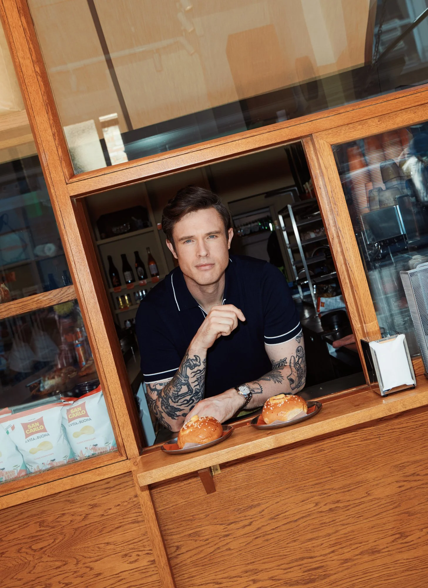 A young man with tattoos on his arms and wearing a black shirt with white trim is looking through a wooden window at the camera. He is sitting at a table with three sandwiches on plates, and the background shows shelves and bottles in a kitchen or ca
