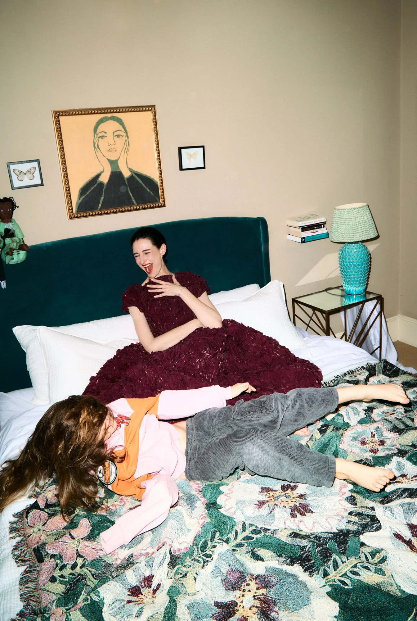 A woman in a maroon dress and a girl in pajamas lying on a bed with a floral quilt. The woman is sitting upright, laughing with her hand on her chest. The girl is lying down, wearing a pink and orange hoodie and gray pants, with long brown hair. Behind them, the wall has three framed artworks, including a large portrait of a woman and two smaller butterfly drawings. There is a teal headboard, a side table with a turquoise lamp, and a small stack of books.