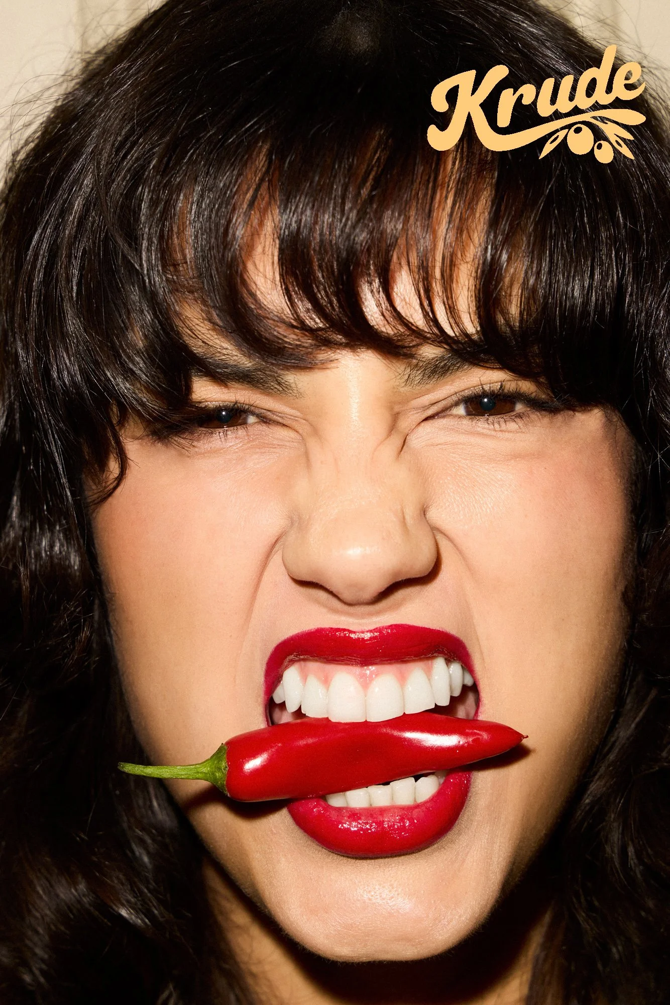 A woman with dark hair and red lipstick biting a red chili pepper with her teeth.