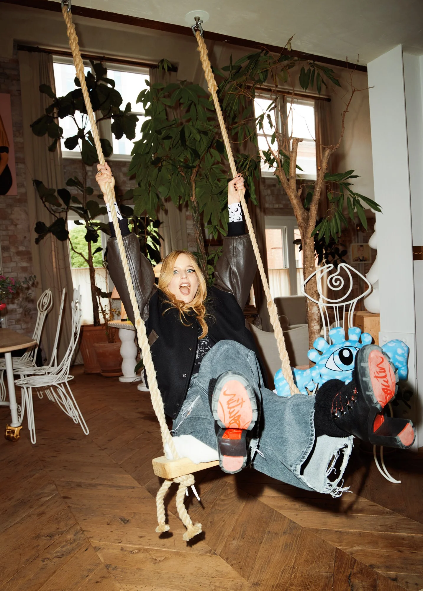 A woman with blonde hair and a black jacket is sitting on a large indoor swing with ropes, in a room decorated with potted plants, wooden floors, and white chairs.