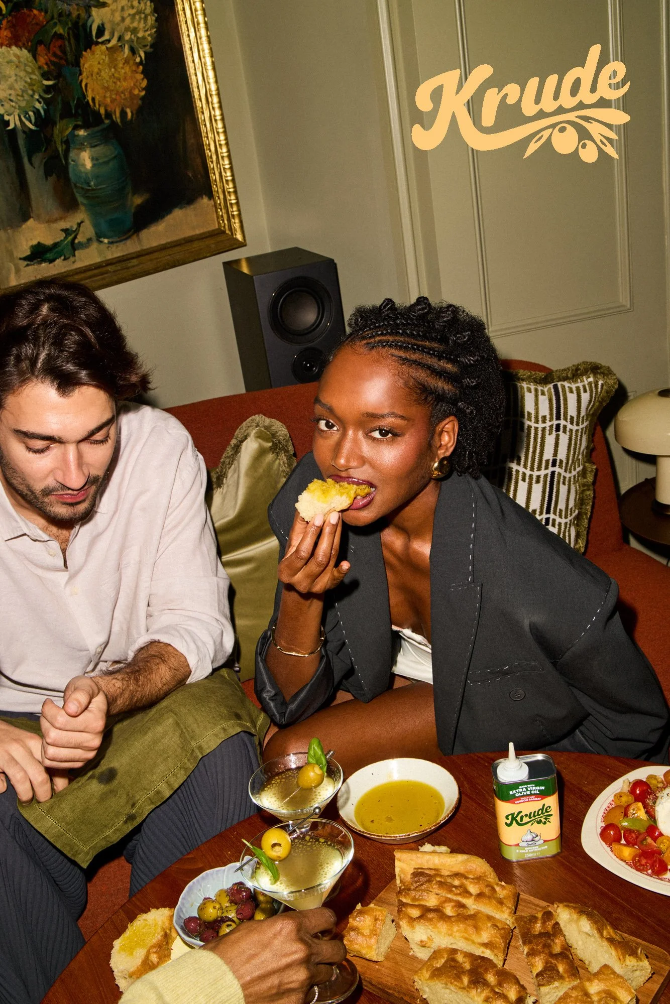 People enjoying a meal around a table with various dishes, bowls of olives, slices of bread, a bowl of olive oil, a plate of cherry tomatoes, and a can of extra virgin olive oil. A woman is eating bread while a man reaches for olives.