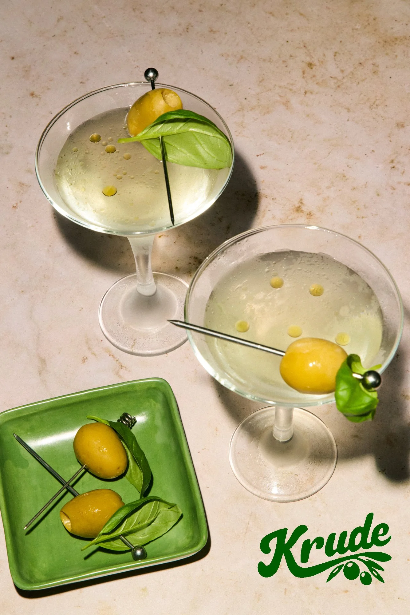 Two glasses of a clear beverage garnished with green basil leaves, yellow olives, and skewers, on a light-colored surface. A green tray with more yellow olives and basil leaves is also visible.