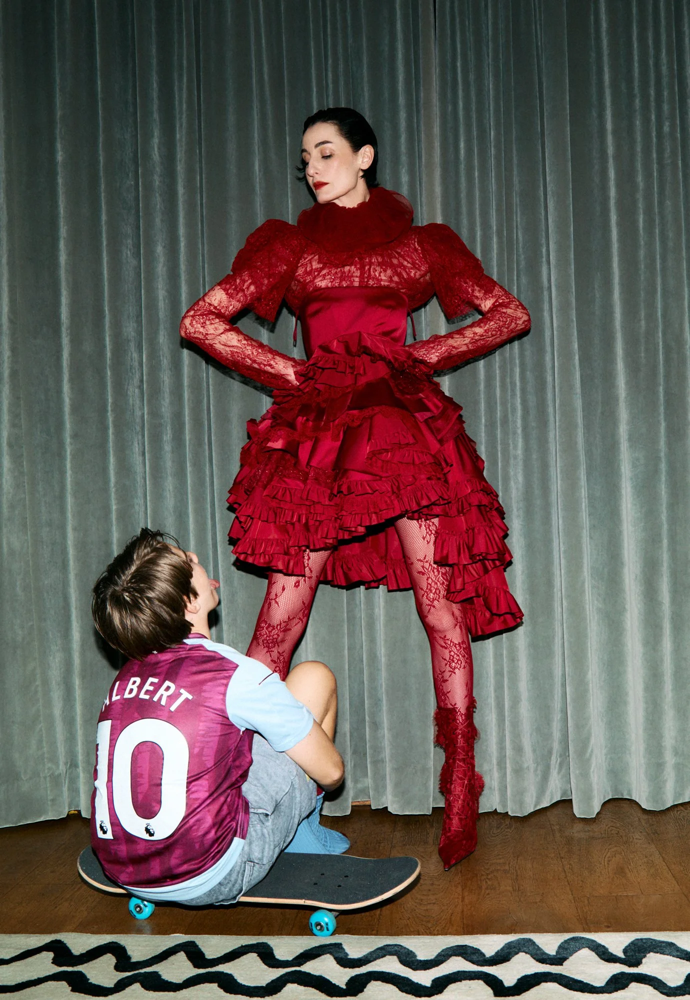 A woman dressed in a red, ruffled dress, standing tall with her hands on her hips, looking down at a boy who is sitting on a skateboard, wearing a sports jersey, and holding up her skirt.