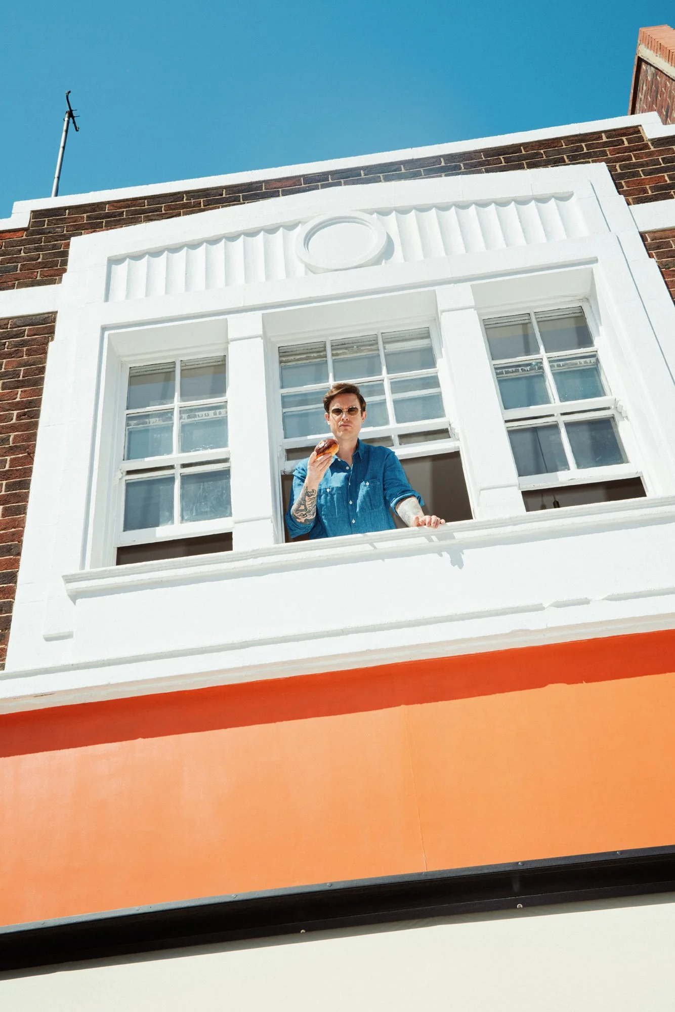 A man with tattoos and sunglasses is standing on a balcony, eating a doughnut, wearing a blue shirt. The building has a white window frame and a brick wall with decorative white plasterwork. The sky is blue and clear.