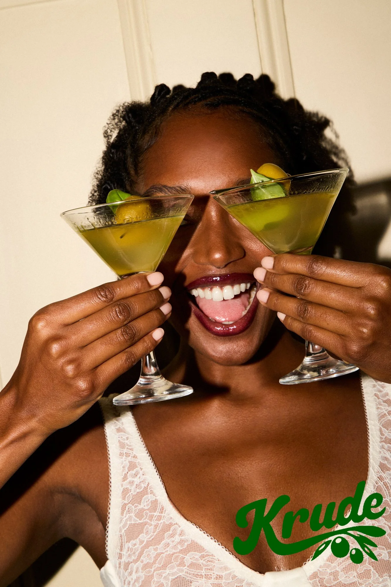 A woman with dark skin, wearing a white lace top with the word 'Krade' in green and an olive branch graphic, holding two martini glasses filled with a yellow-green drink and smiling with her mouth open, holding the glasses in front of her eyes.