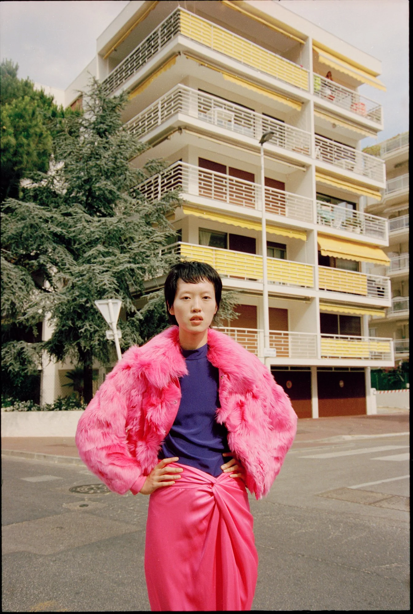 A woman with short black hair, wearing a pink faux fur jacket and a bright pink skirt, poses outdoors with her hands on her hips in front of a mid-century modern apartment building with yellow and white balconies and trees.