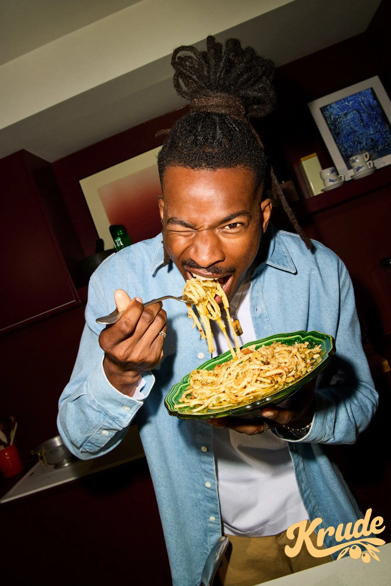 A man with dreadlocks piled on his head, wearing a light blue denim shirt and tan pants, is eating a plate of fettuccine pasta with a fork, smiling and winking at the camera, in a kitchen with dark wood cabinets and decorative cups in the background.
