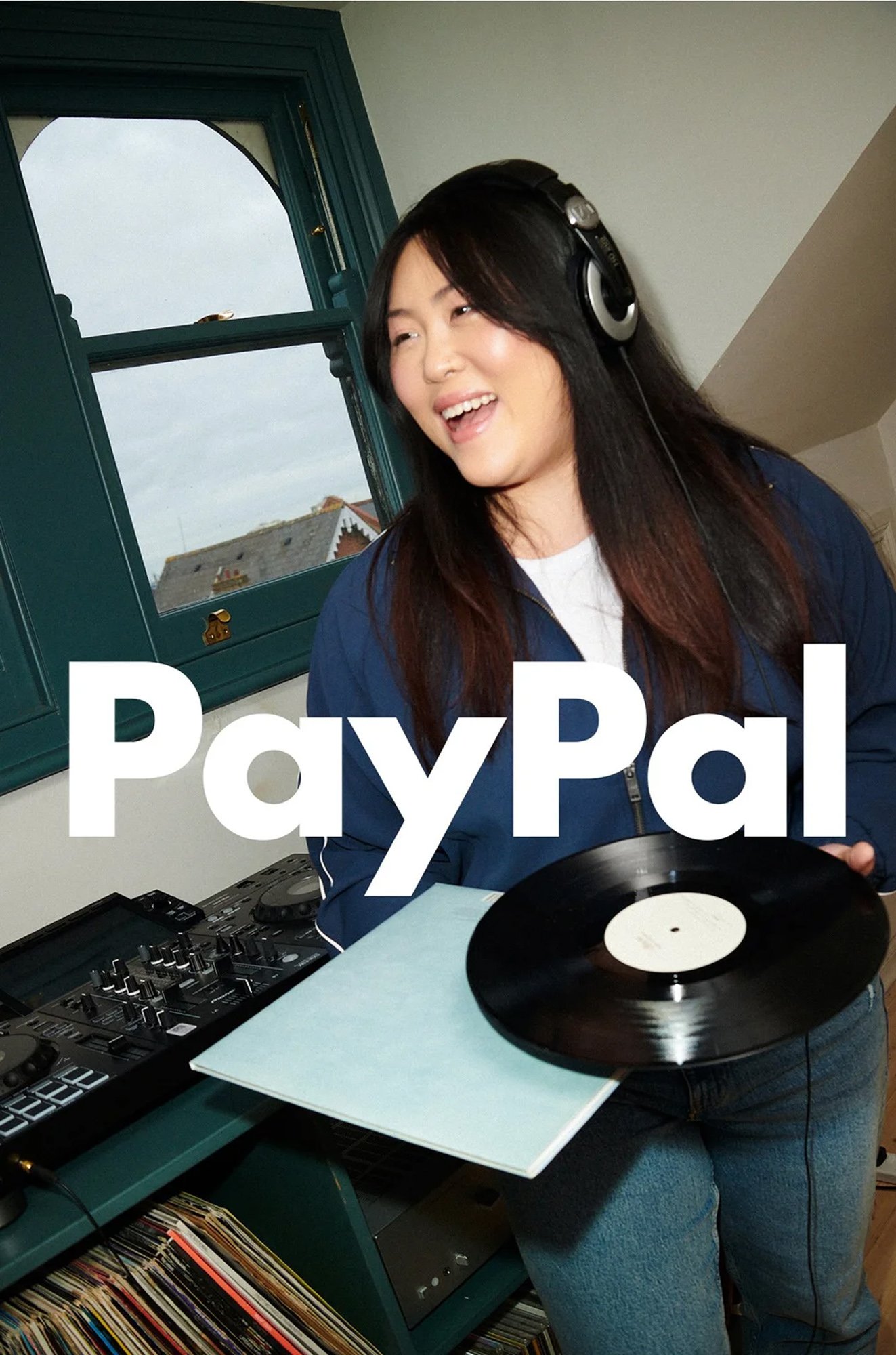 A woman with long dark hair wearing headphones, standing in front of DJ equipment and a vinyl record, with the PayPal logo overlaid on the image.