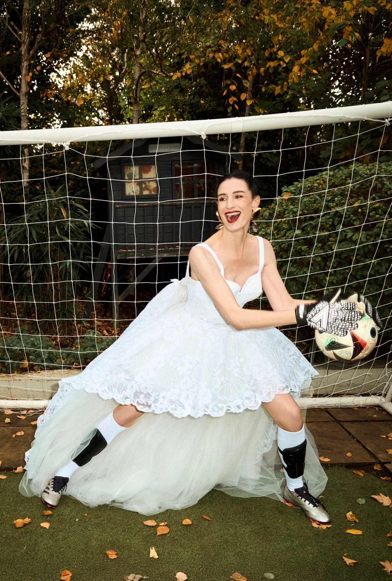 A woman dressed in a white wedding gown and soccer gear, including cleats, knee pads, and goalkeeper gloves, is standing in a soccer goal area holding a soccer ball with a smile. The background features a net, trees, and autumn leaves on the ground.