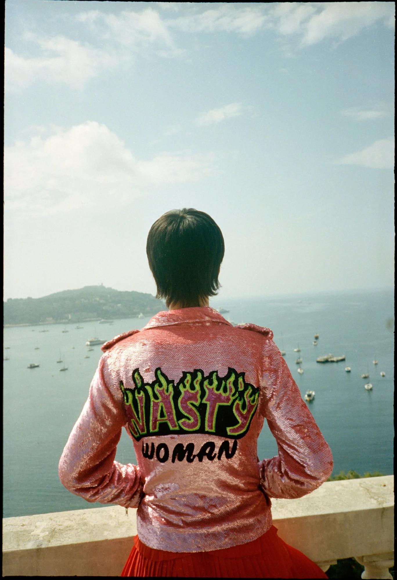 Person with short hair wearing a pink, shiny, embroidered jacket that says 'Nasty Woman' with flames, standing on a balcony overlooking a harbor with boats, hills, and cloudy sky in the background.