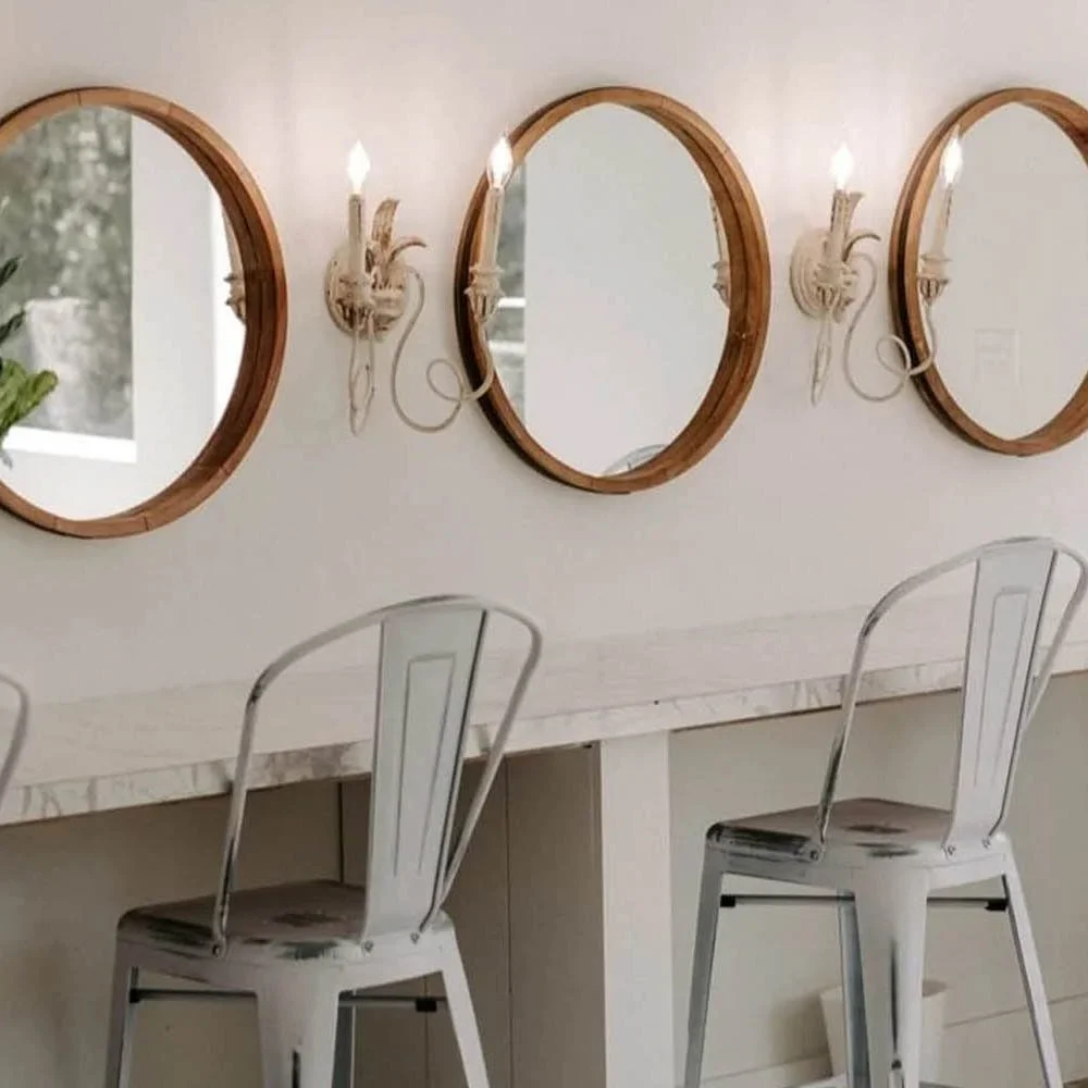 A row of three round mirrors with wooden frames on a white wall, each flanked by two wall-mounted candle sconces with lit candles. In front is a white marble-topped counter with two white metal bar stools.