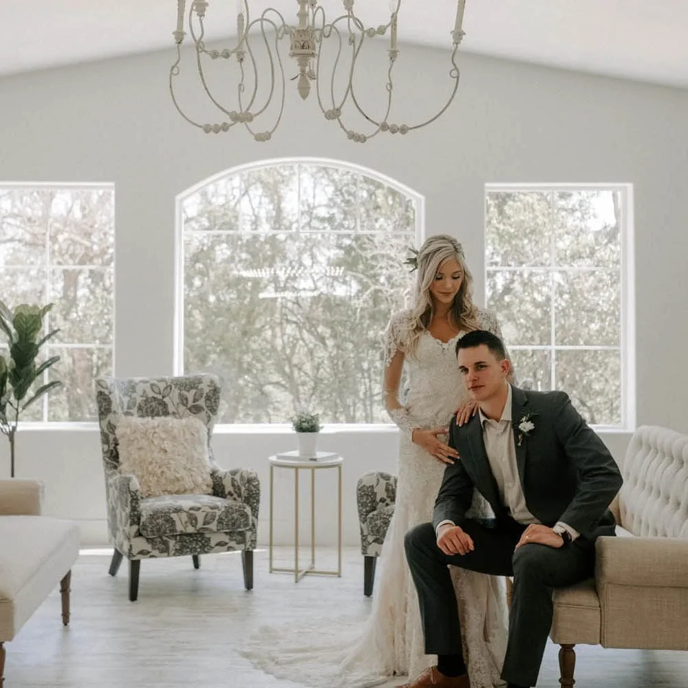 A bride and groom in a bright, elegant room with large windows, a chandelier, and vintage-style chairs.