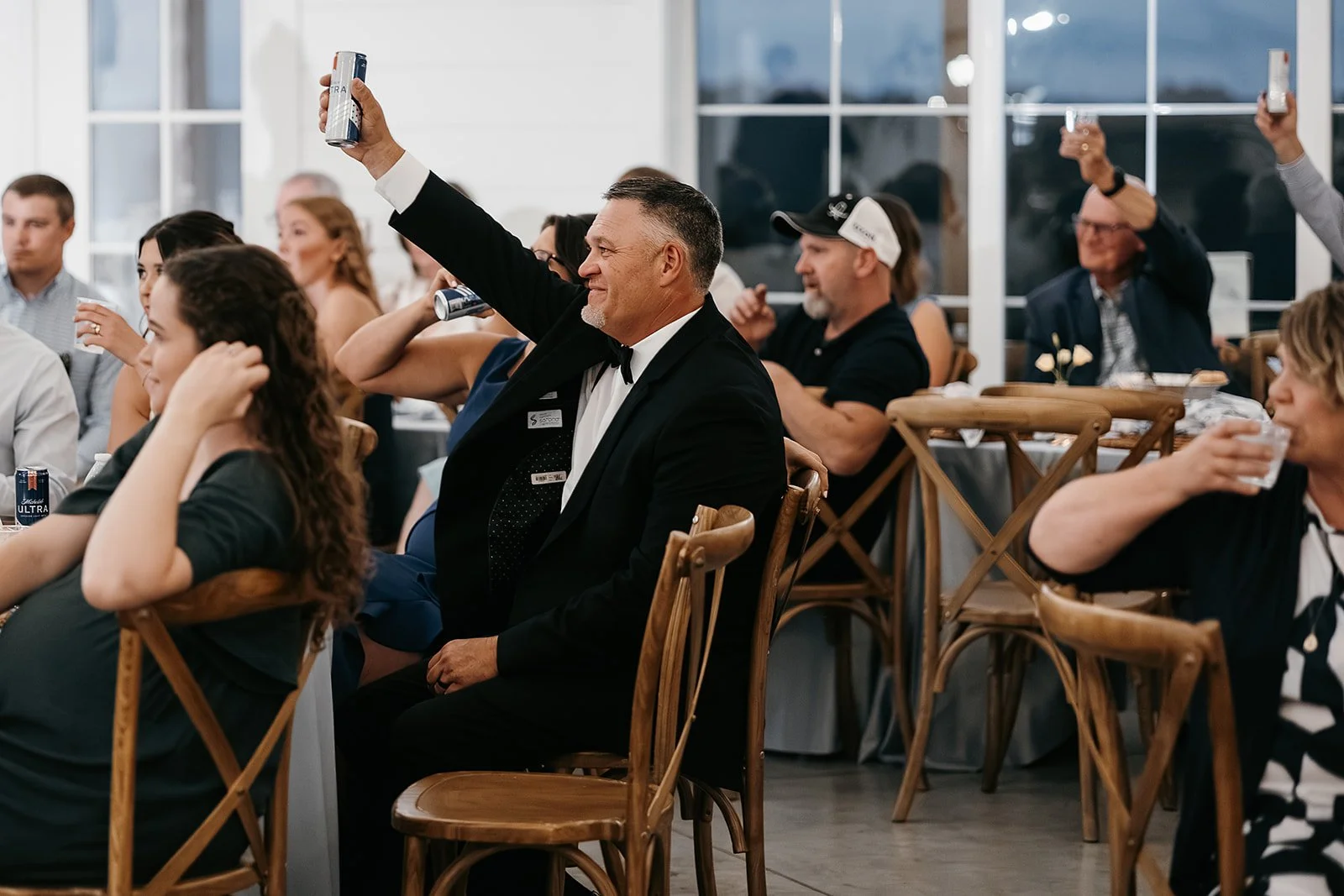 People in formal and casual attire seated at tables in a brightly lit room, raising cans and glasses in a toast or celebration.