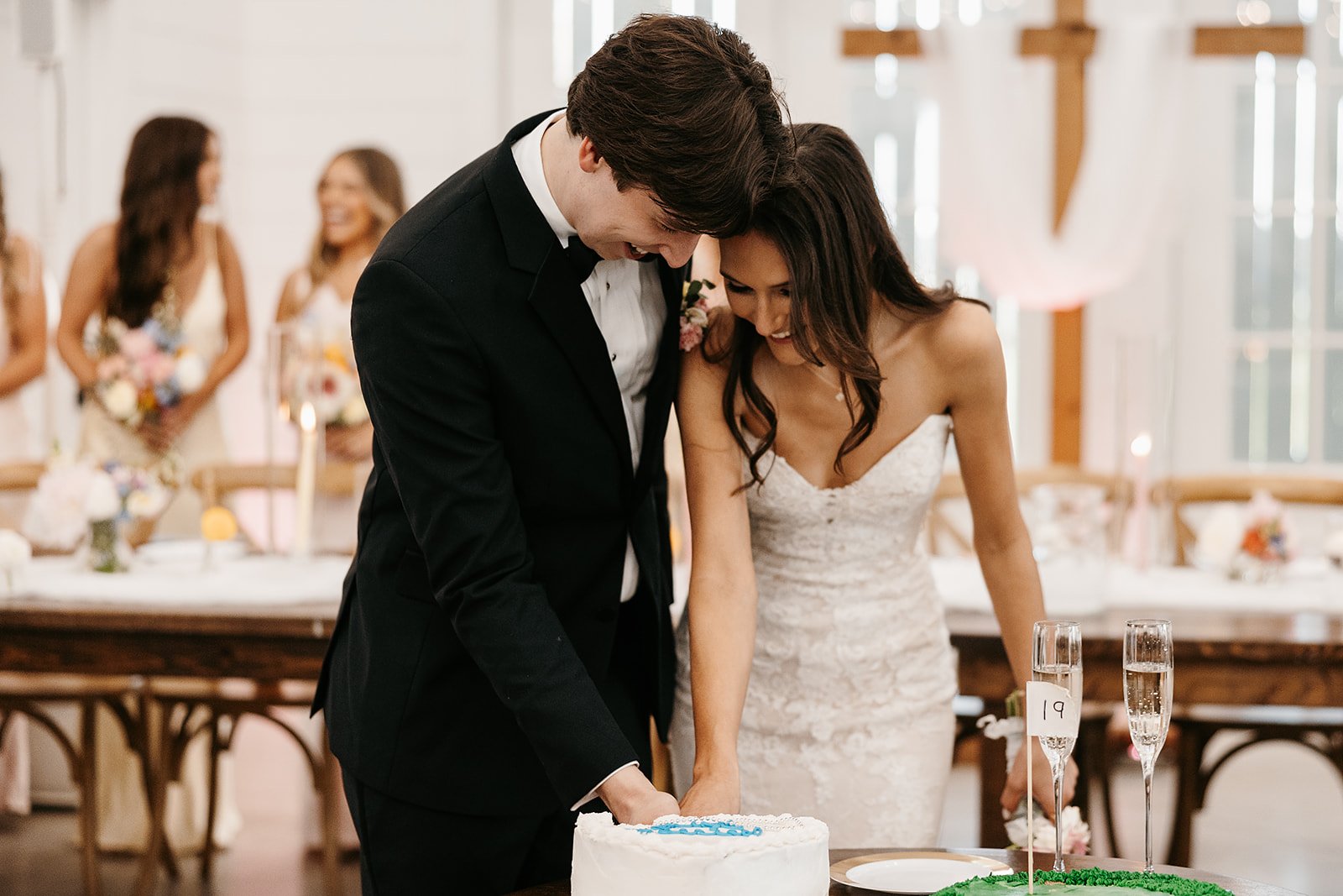 Bride and groom cut a wedding cake at their reception, with bridesmaids in the background.