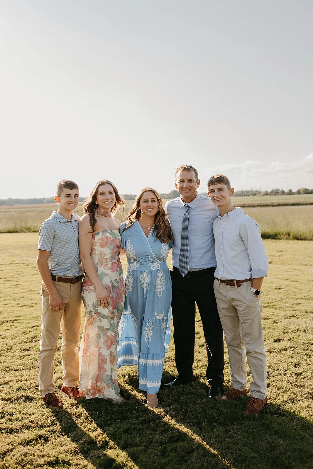 A group of six people standing outdoors in a field, smiling at the camera during late afternoon or early evening. There are three women and three men, dressed in casual or semi-formal clothing, with a clear sky and open landscape in the background.