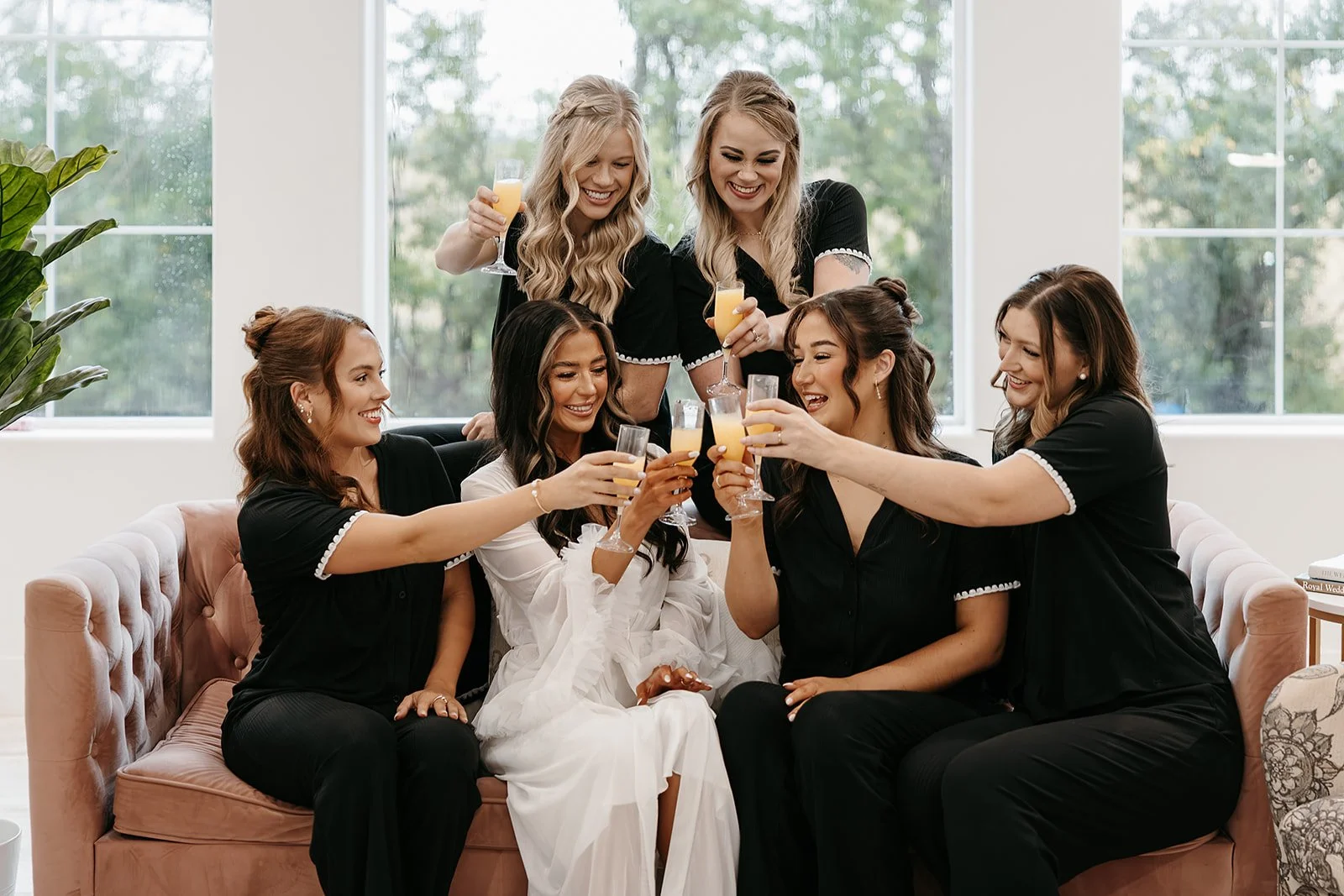 bride with her bridal party on pink couch toasting mimosas