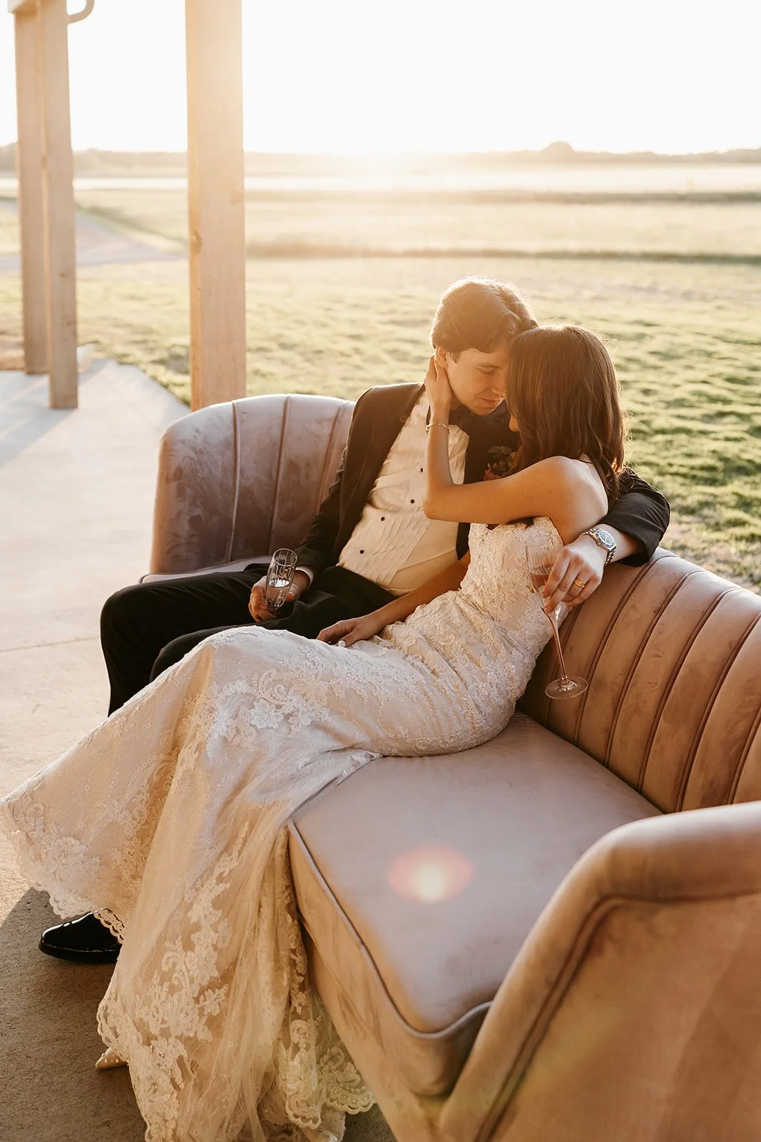 A newlywed couple sitting on a vintage sofa outdoors at sunset, with the bride in a lace wedding gown and the groom in a tuxedo, sharing an intimate moment with champagne glasses.