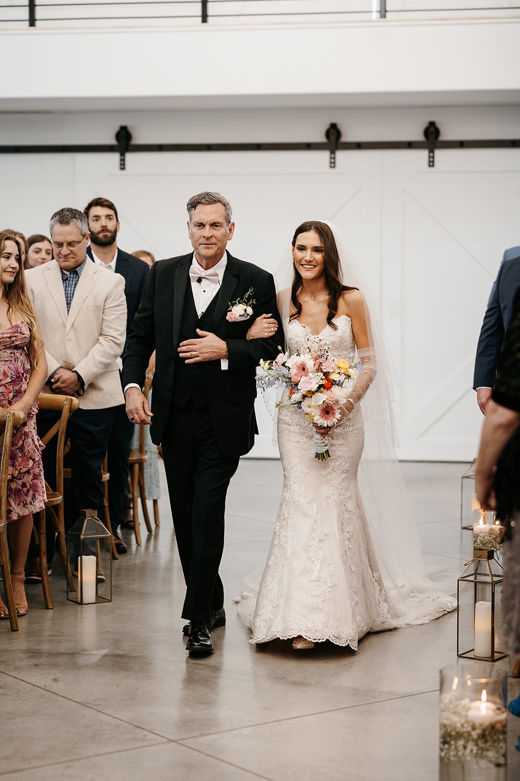 Bride walking down the aisle in a wedding ceremony, accompanied by a man in a tuxedo, inside a decorated venue with candles and lanterns.