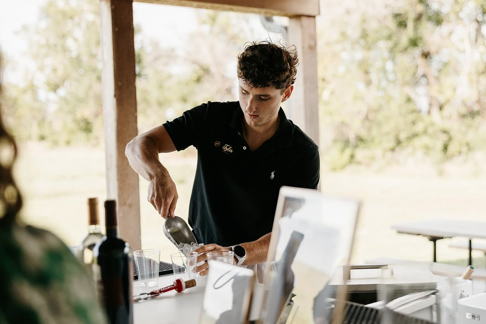 A young man with curly brown hair wearing a black polo shirt with a white logo, pouring a liquid into a glass at an outdoor event or gathering under a wooden structure, with bottles and other items on the table.