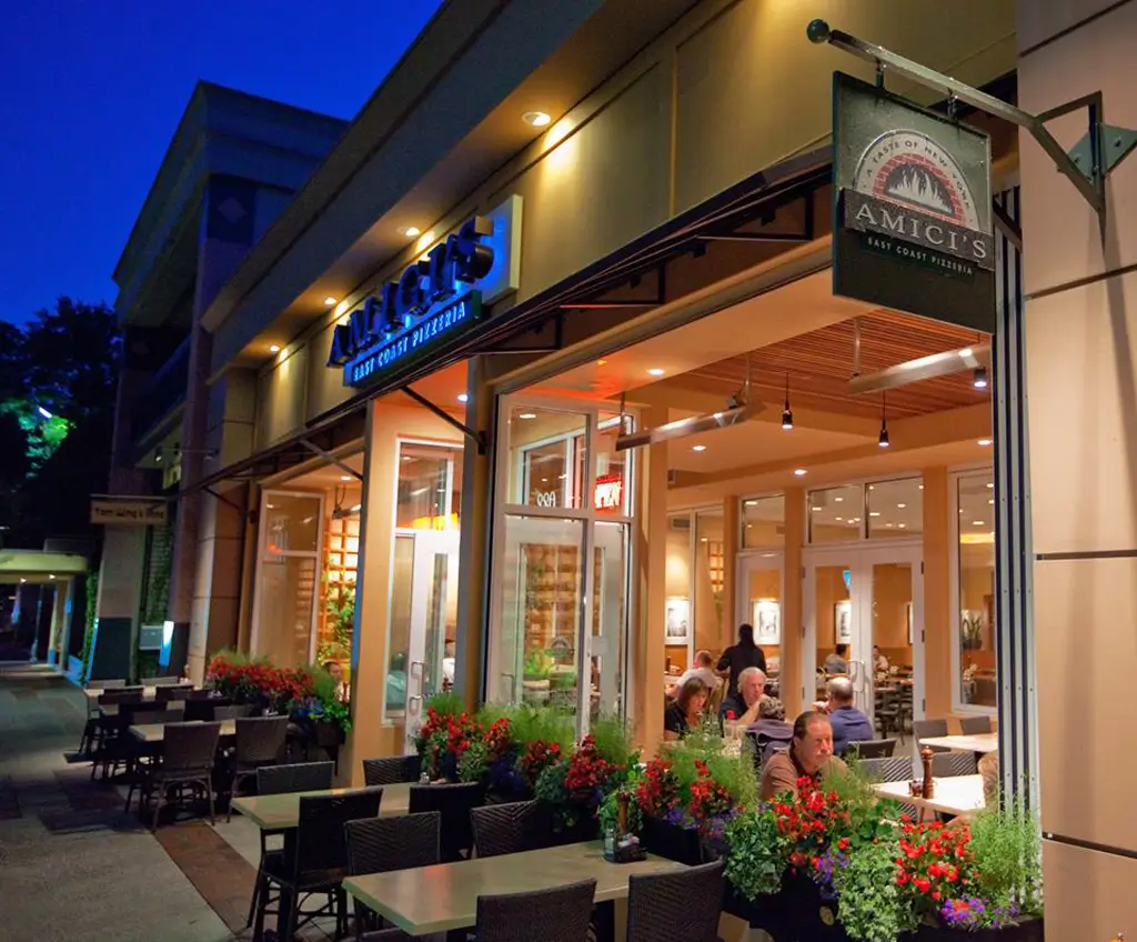 A warmly lit Amici’s East Coast Pizzeria storefront at night with people dining on the Menlo Park patio.