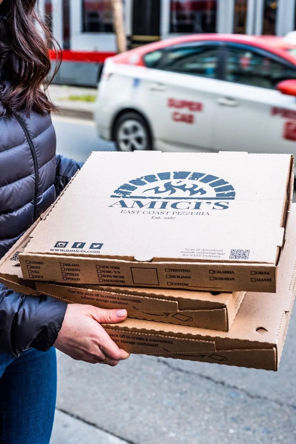 Delivery driver carrying three stacked Amici’s East Coast Pizzeria pizza boxes while making a food delivery in the city.