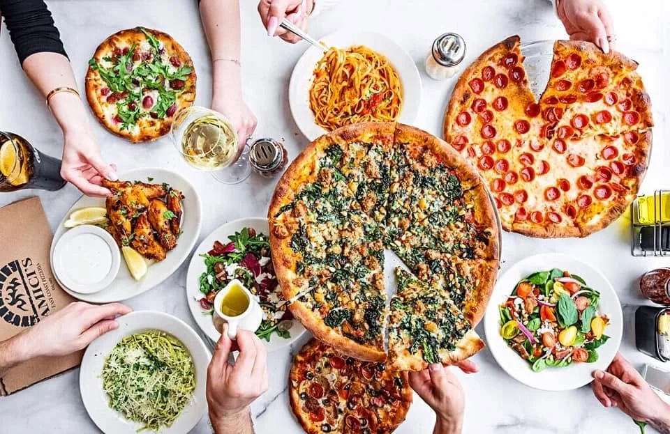 Colleagues sharing an Amici's catering spread with pizza, pasta, and fresh salads at an office gathering.